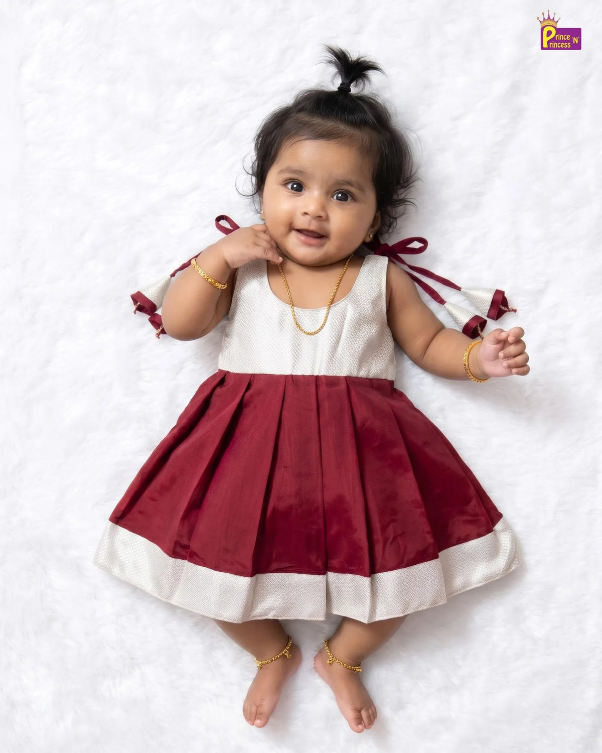 A baby girl wearing a sleeveless maroon and white dress with pleated skirt and bow details, lying on a white textured blanket.