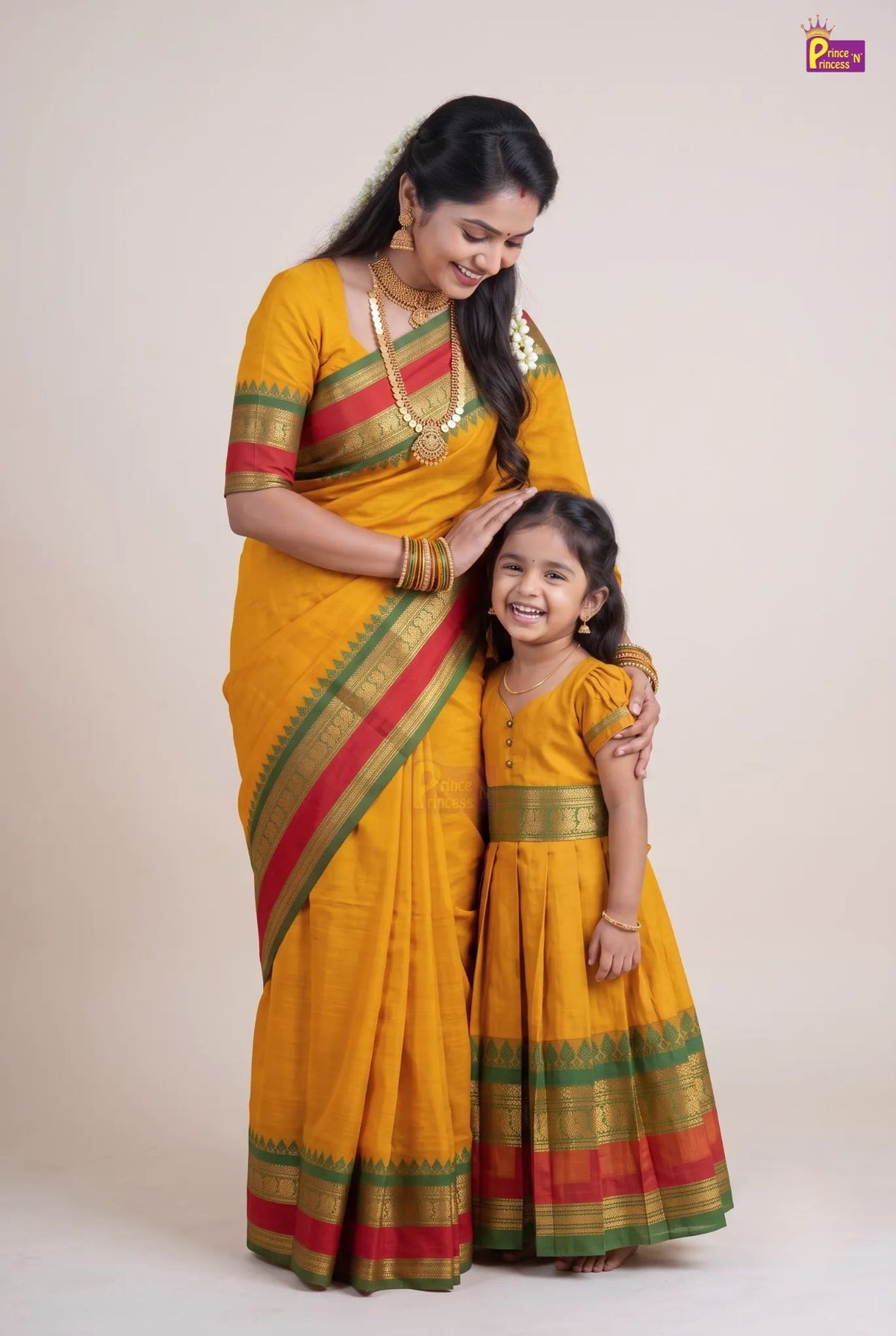 A woman and a young girl wearing matching mustard yellow traditional Indian outfits with red and green striped borders, standing against a plain beige studio background.