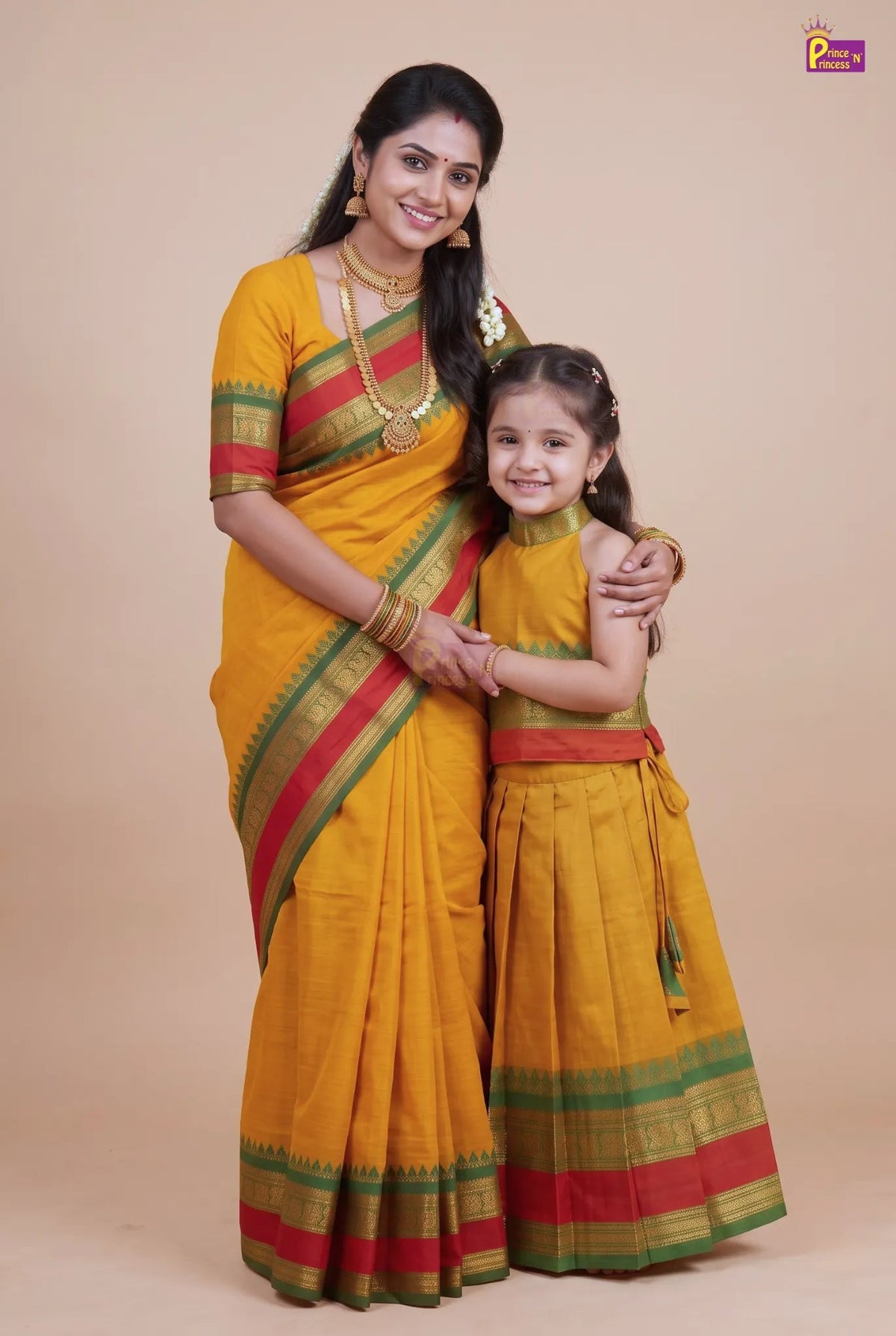 A woman and a young girl posing together in matching traditional Indian attire. Both are wearing yellow sarees and matching long kurta sets with green and red borders, set against a solid beige studio background.