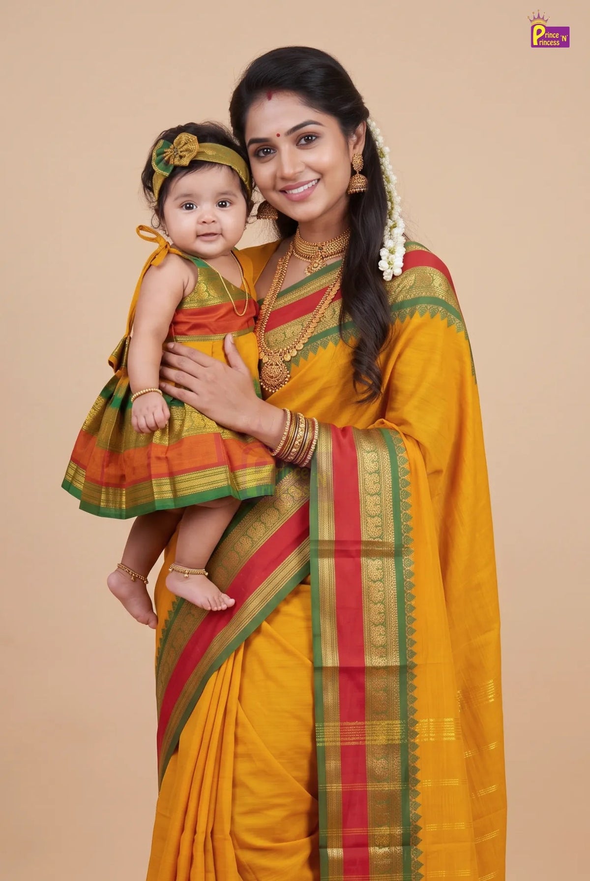 A woman and a baby wearing matching traditional Indian attire, featuring a mustard yellow saree with red and green borders, posed against a solid beige studio background.