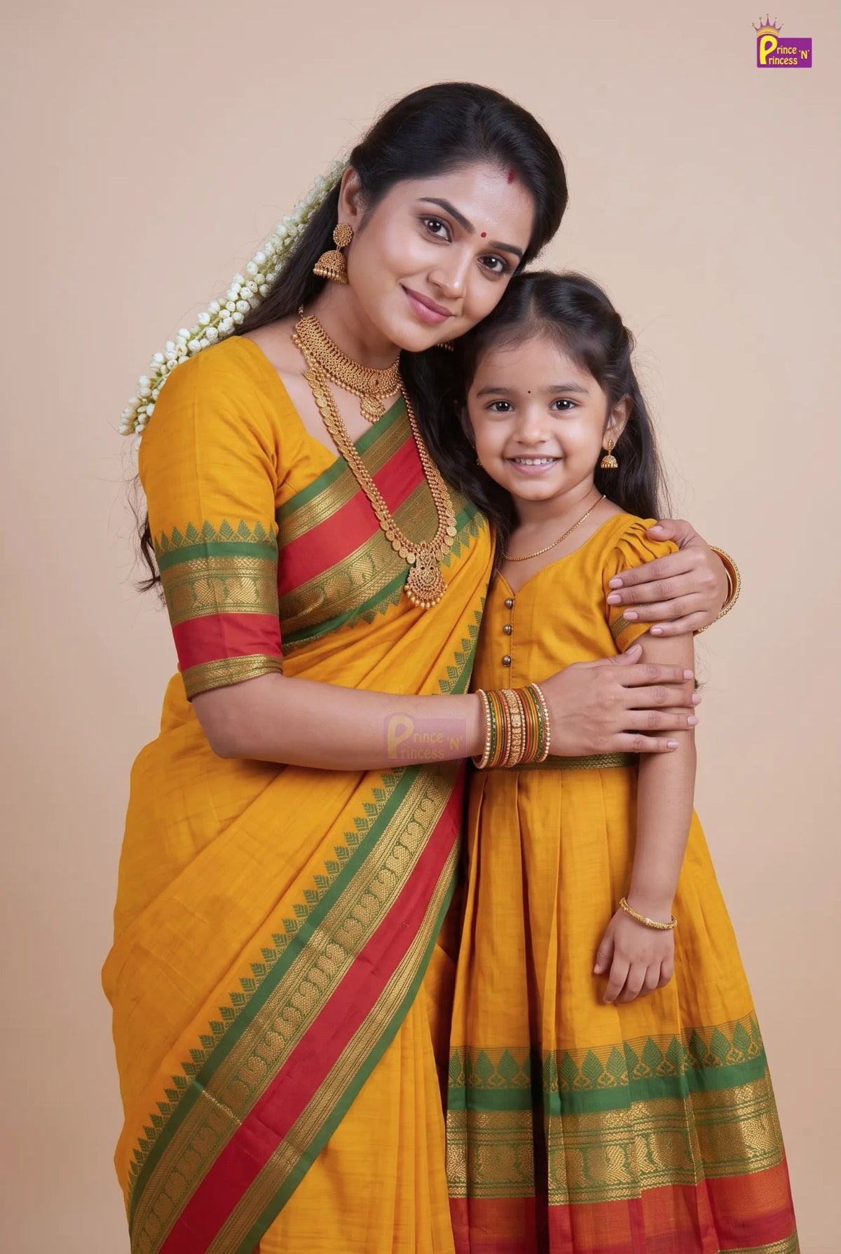 A woman and a young girl wearing matching yellow traditional Indian outfits with red and green borders, standing against a plain beige studio background.
