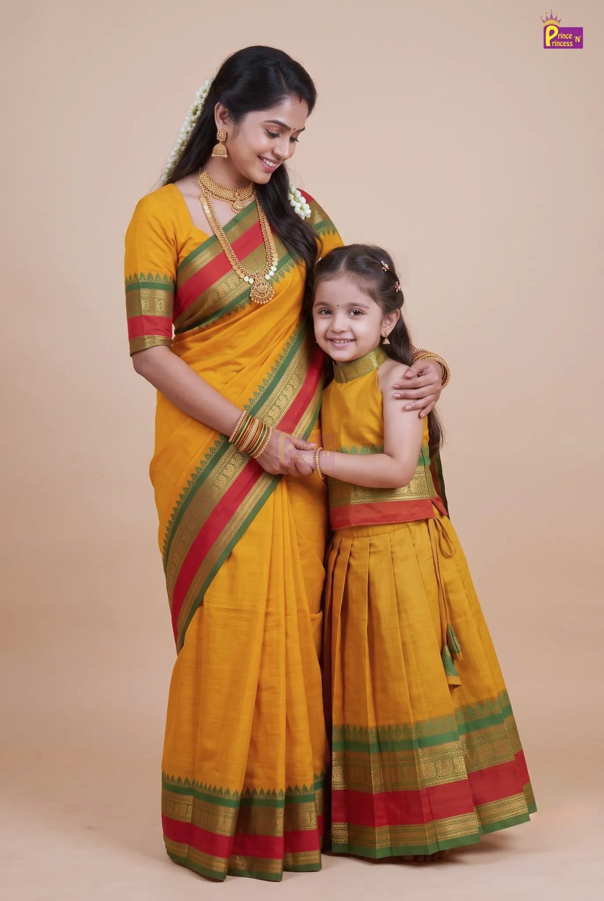 A woman and a young girl wearing matching yellow traditional Indian saris with green and red borders, standing together against a plain beige studio background.