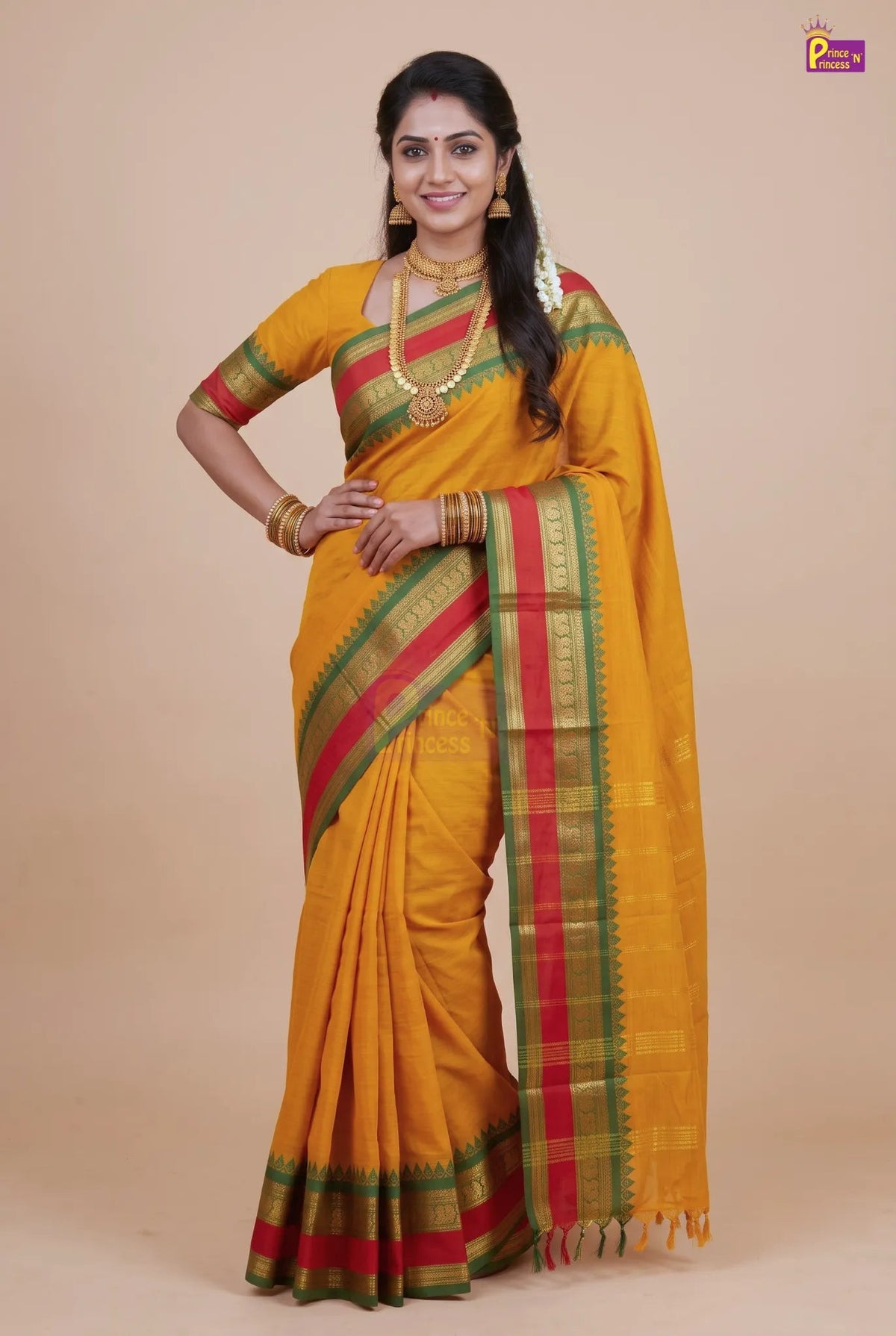 A woman modeling a yellow silk saree with red and green borders, paired with gold jewelry, against a solid beige background.