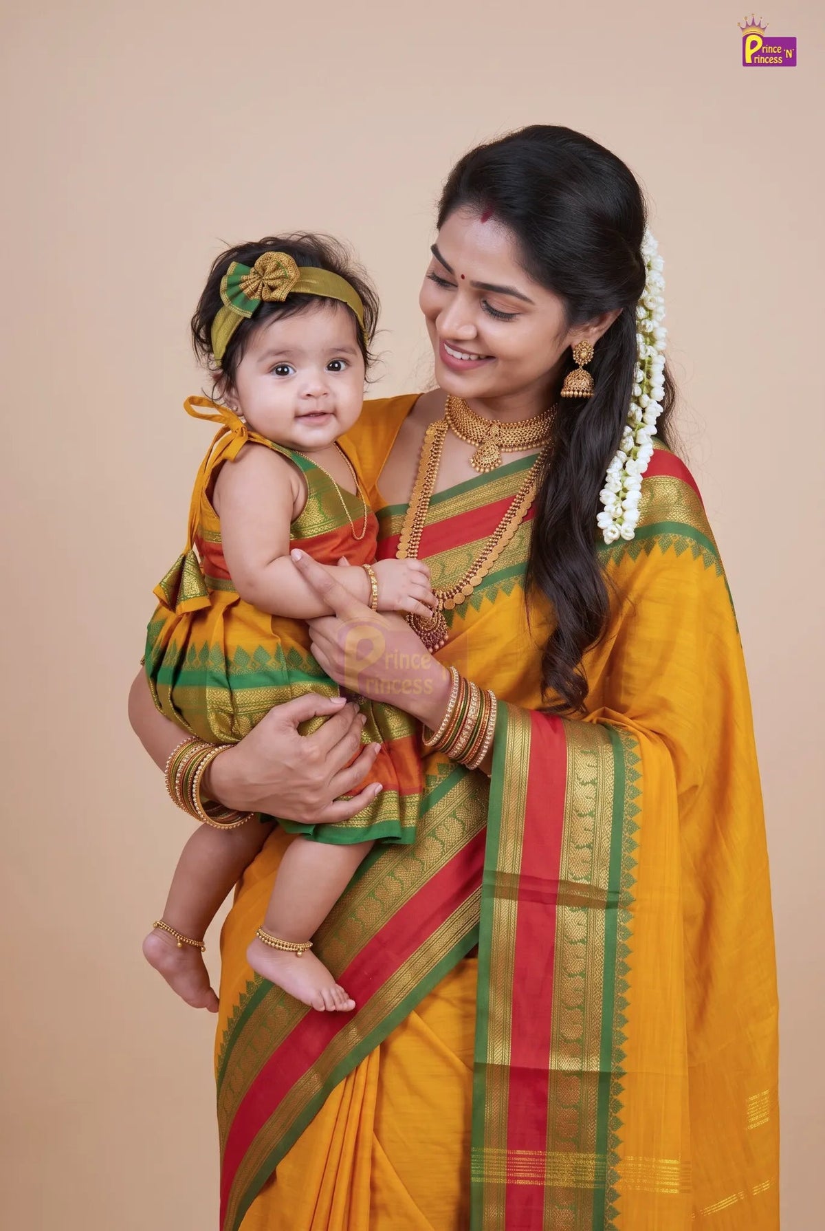 A woman and a baby wearing matching traditional Indian outfits, featuring a mustard yellow, green, and red saree and dress, posed against a solid beige studio background.