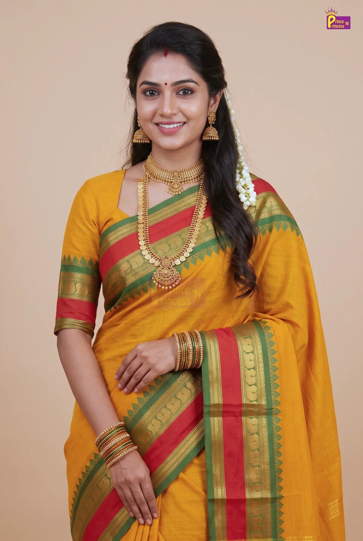 A woman wearing a yellow silk saree with red and green stripes, gold jewelry, and traditional accessories, posed against a solid beige background.