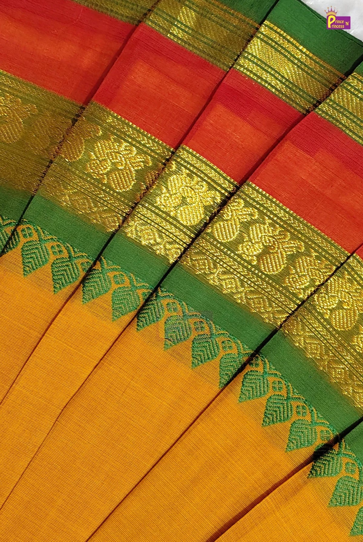 A close-up of a folded traditional fabric, likely a saree, featuring a mustard yellow body, a red-orange upper section, and a green border with intricate gold zari woven patterns.