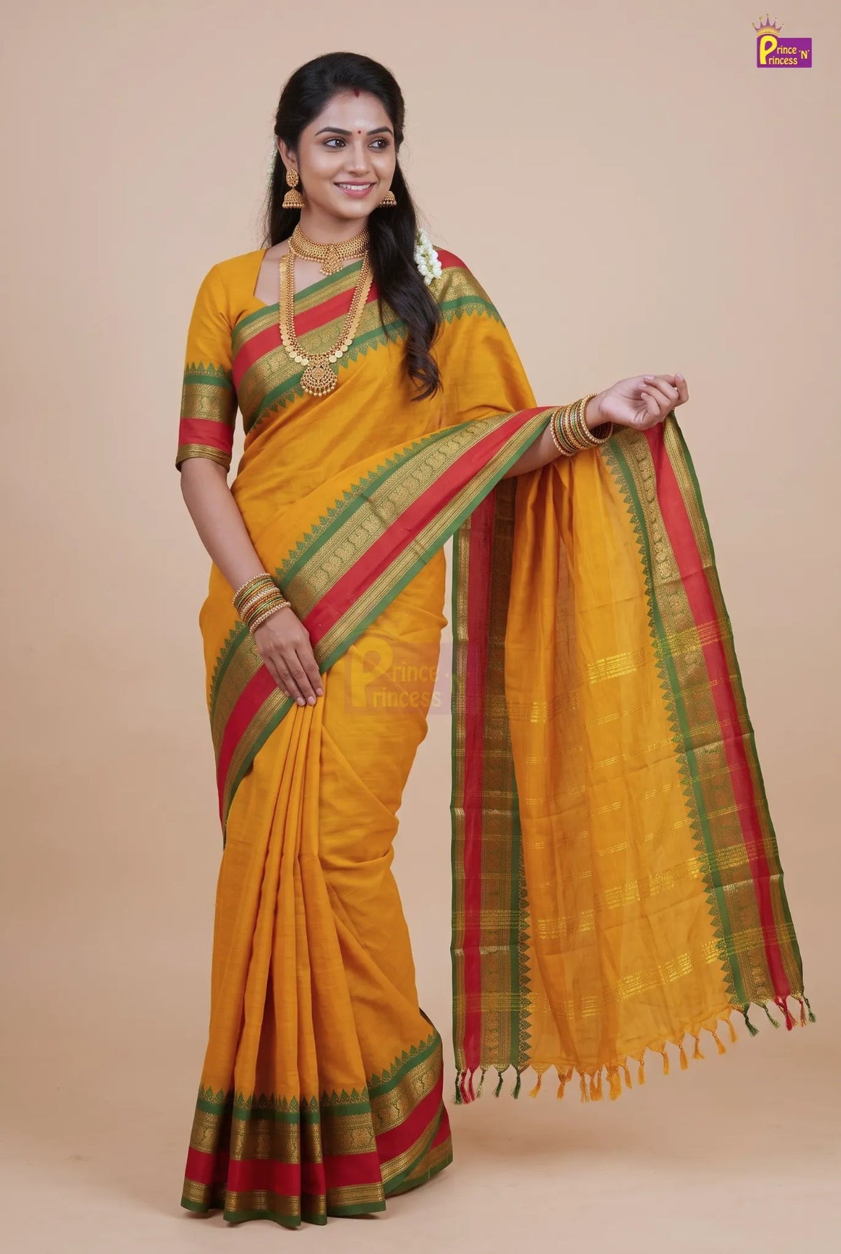 A woman modeling a traditional Indian saree in mustard yellow with green and red borders, styled with gold jewelry against a solid beige background.