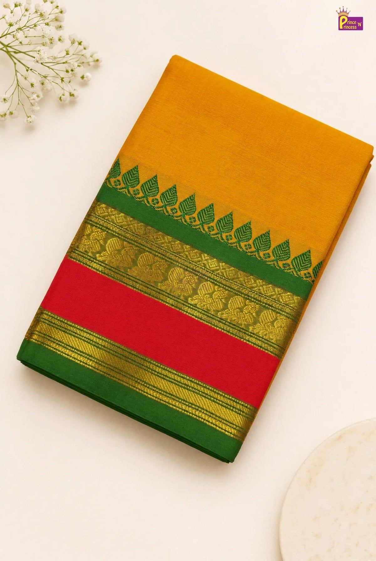A folded yellow-orange silk saree with a green border, gold zari detailing, and a wide red band, displayed on a light cream background with decorative baby's breath flowers.