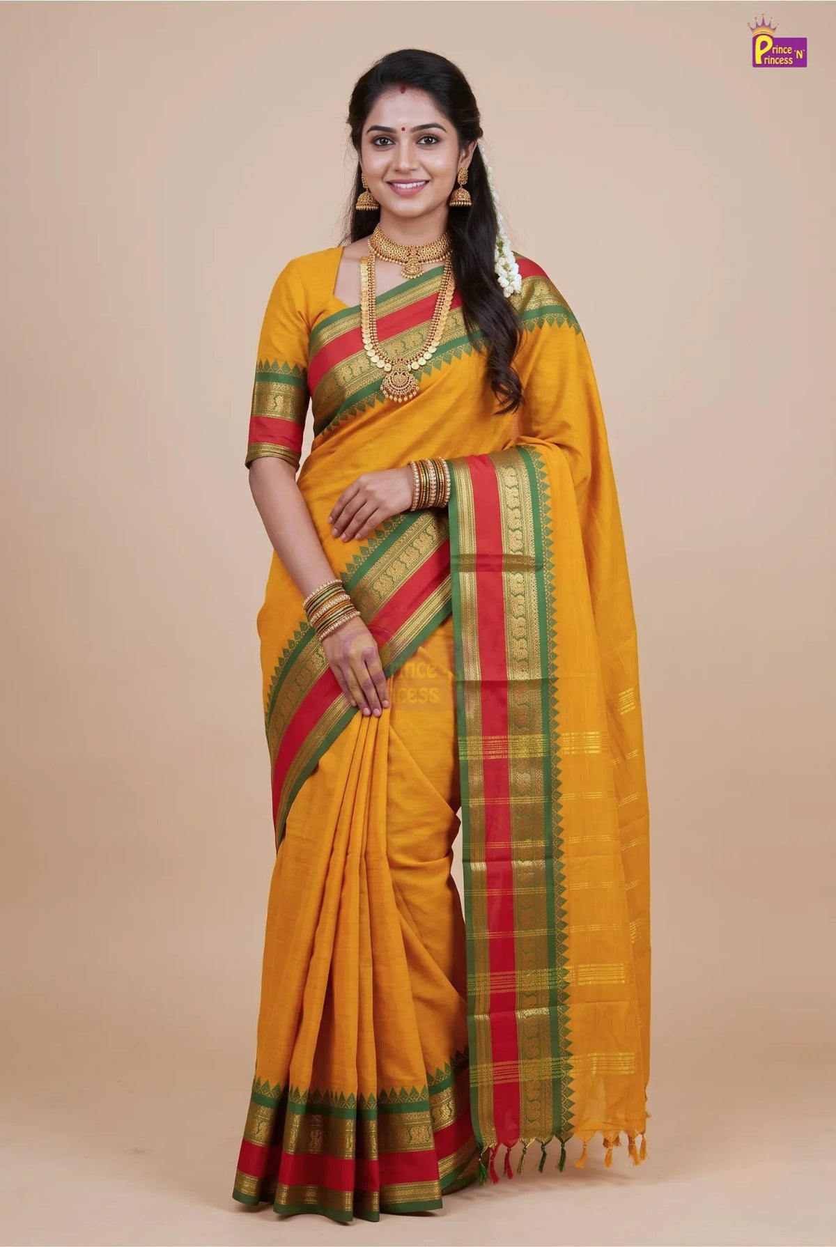 A woman modeling a traditional Indian saree in mustard yellow with red and green borders, paired with a matching yellow blouse. She is standing against a solid beige background.