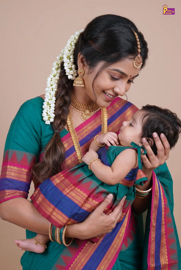 A woman in a traditional green saree with blue, pink, and gold stripes holds a baby wearing a matching green outfit. The woman has white floral hair ornaments and gold jewelry, and the baby has a gold headpiece.