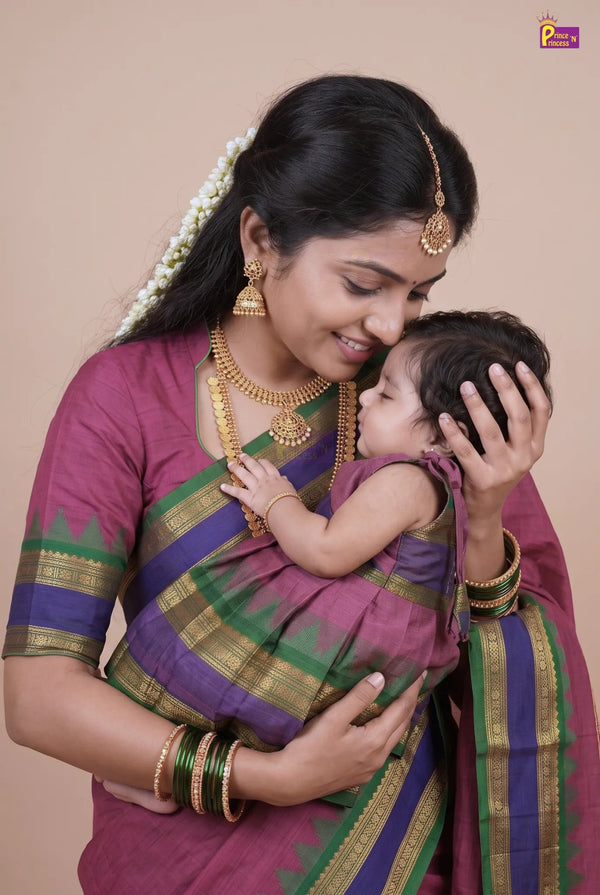 A woman in traditional Indian attire wearing a purple, green, and gold sari holds a baby wrapped in a matching purple and green sari. The woman is adorned with gold jewelry, including a maang tikka, earrings, and necklaces, and wears green bangles.