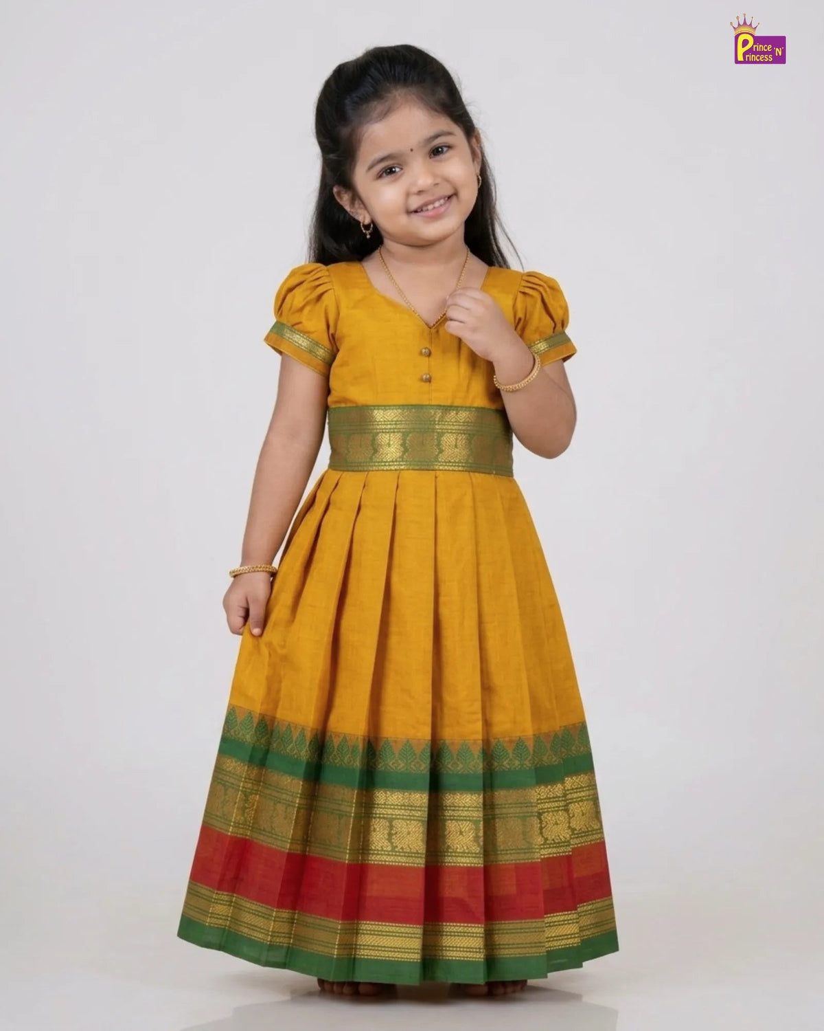 A young girl modeling a traditional Indian yellow cotton dress with green and red borders and gold zari detailing, set against a plain light gray background.