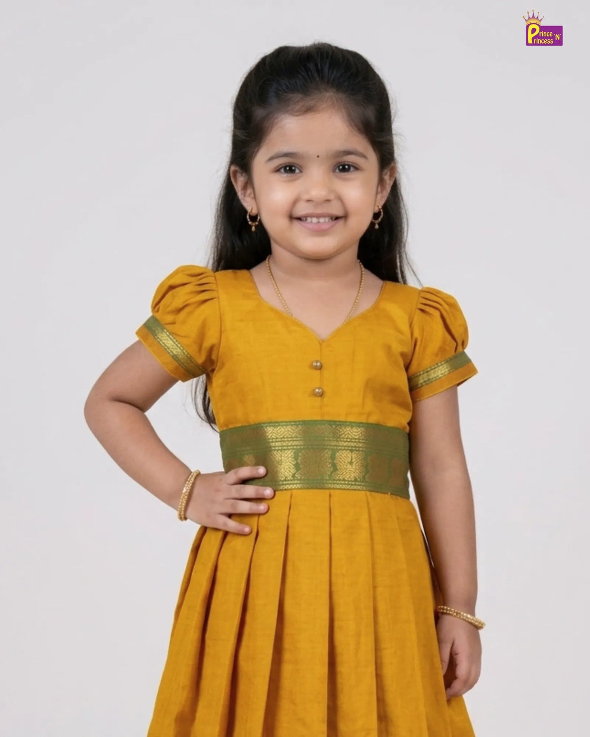 A young girl with dark hair modeling a mustard yellow traditional dress with gold trim and a decorative waistband, set against a plain light gray background.