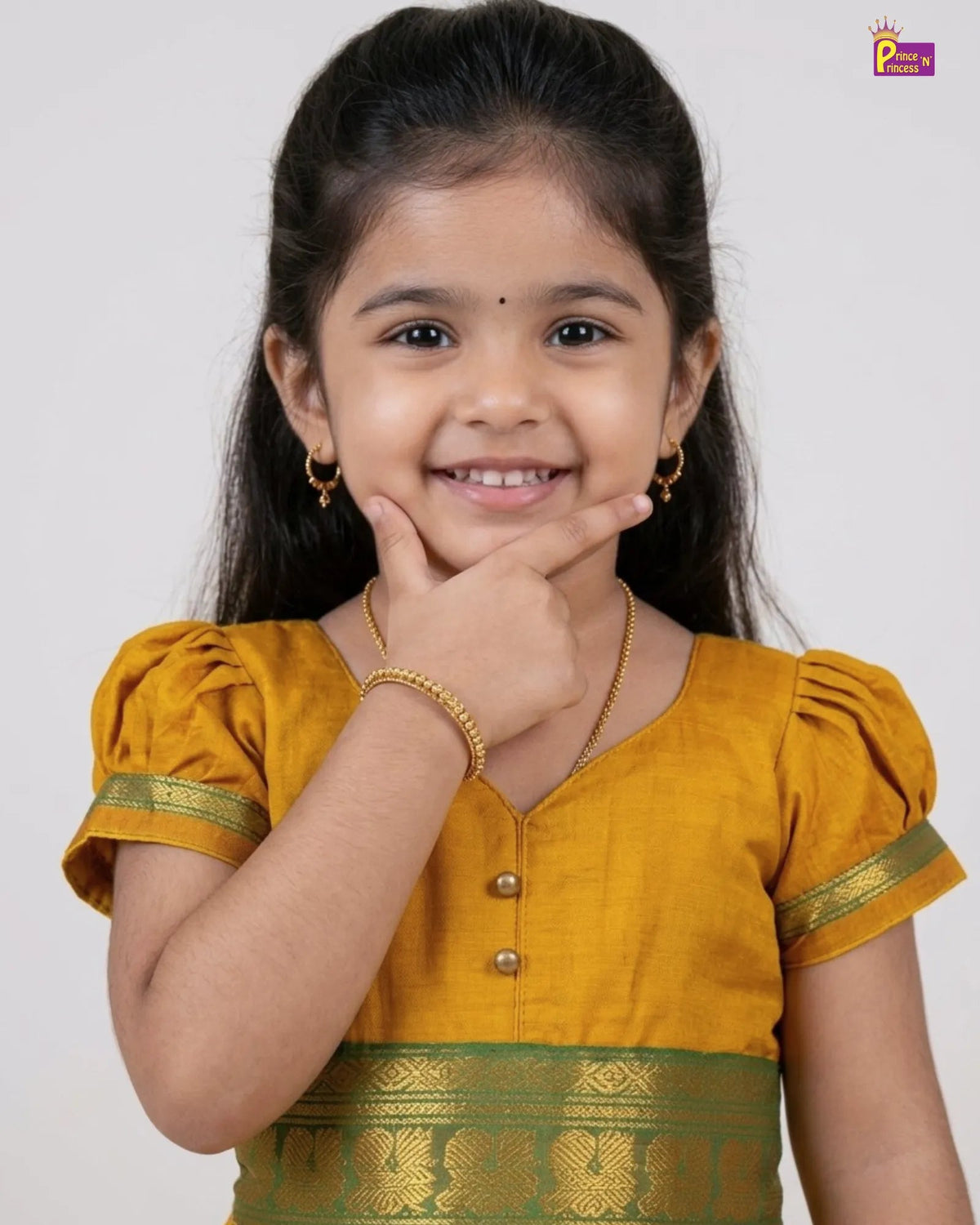 A young girl smiling and posing in a traditional Indian outfit. She is wearing a mustard yellow blouse with gold trim and a matching yellow-gold skirt with a green and gold border, accessorized with gold jewelry against a plain light gray background.