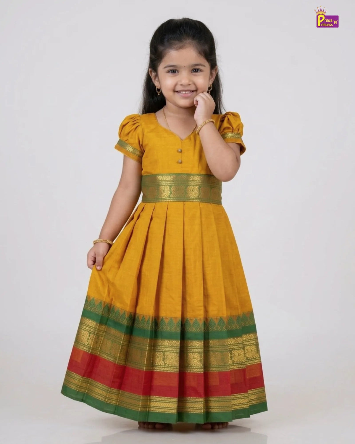 A young girl modeling a traditional Indian yellow cotton dress with green and red borders and gold zari detailing, set against a plain light gray background.