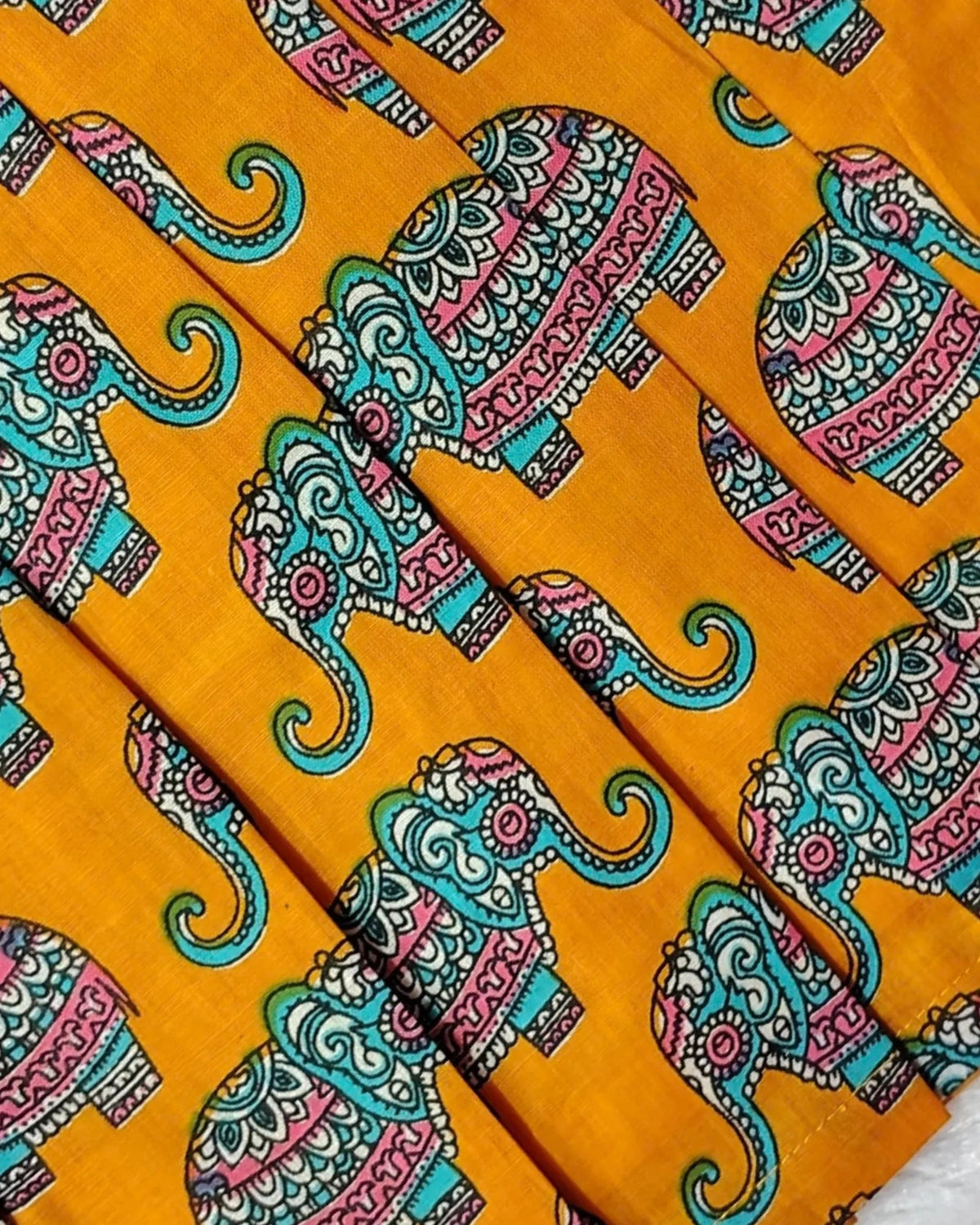 A close-up of a folded fabric with a vibrant mustard yellow background and a repeating pattern of stylized elephants decorated with teal, pink, and white intricate designs.
