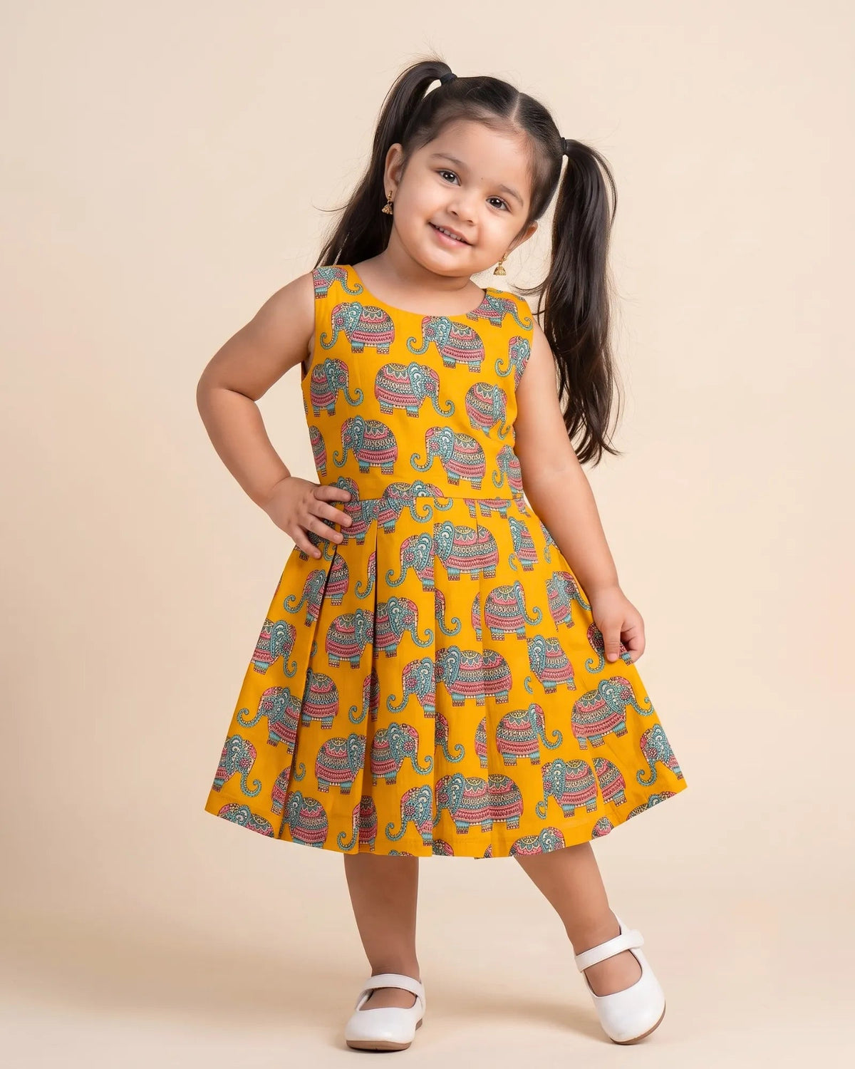 A young girl with dark hair in pigtails stands against a solid beige background wearing a sleeveless mustard yellow dress with a colorful elephant print and white Mary Jane shoes.