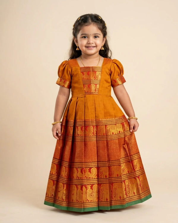 A young girl modeling a traditional Indian dress in orange and gold with a green border, set against a solid beige background.