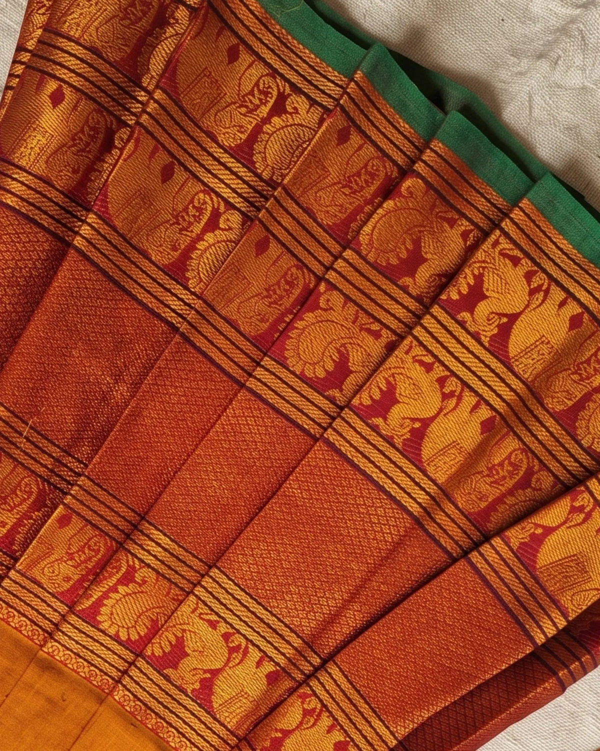 A close-up of a folded traditional Indian silk saree, showcasing its rich rust-orange body, golden zari woven motifs, and a contrasting green border.