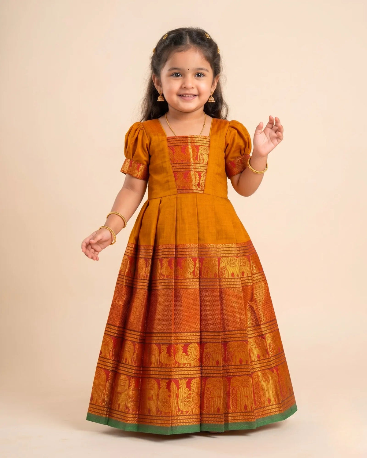 A young girl modeling a traditional Indian dress in mustard yellow and orange with gold zari embroidery and a green border, set against a solid beige background.
