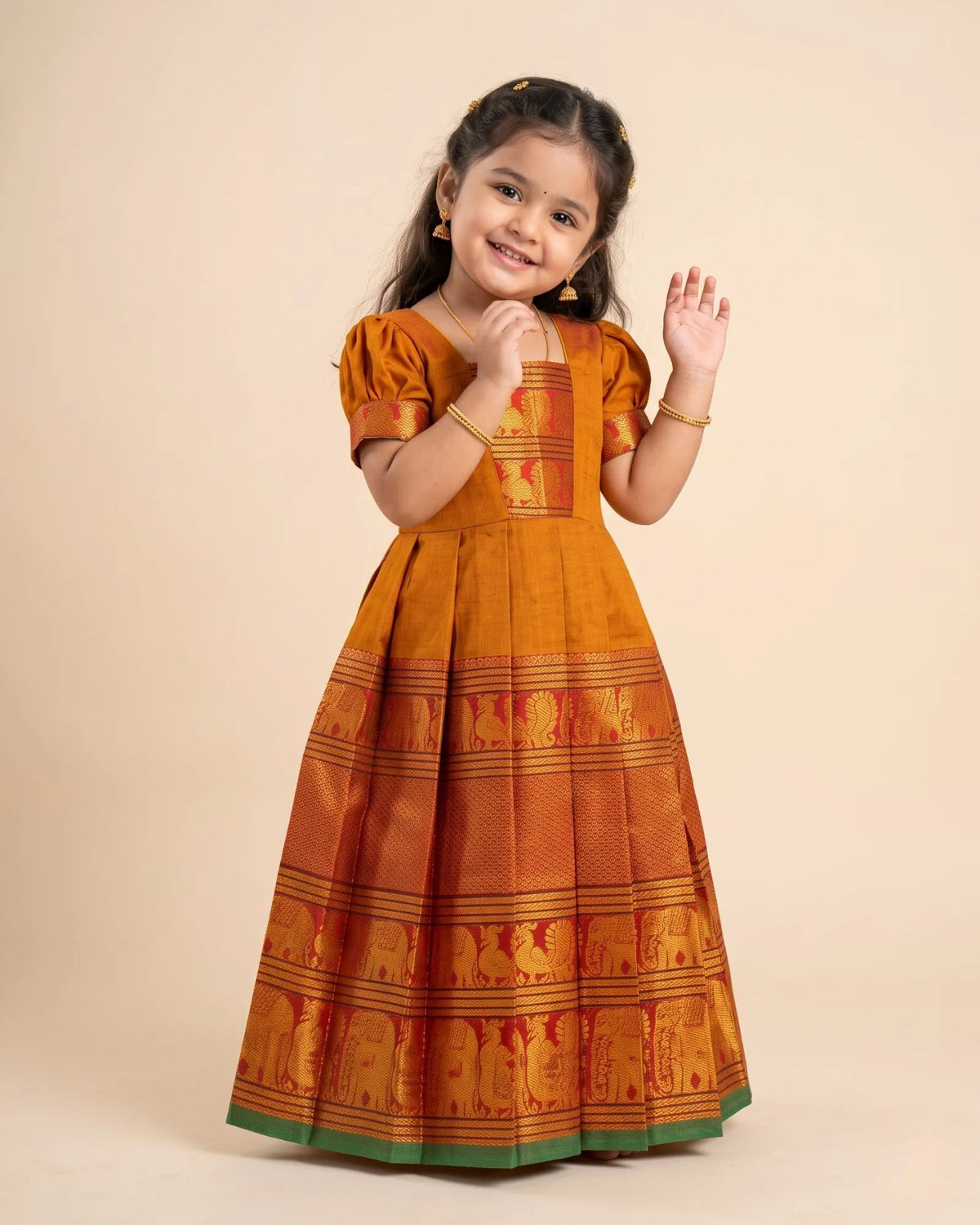 A young girl modeling a traditional Indian Anarkali gown in mustard yellow and orange with red and green borders featuring elephant motifs, set against a solid beige studio background.