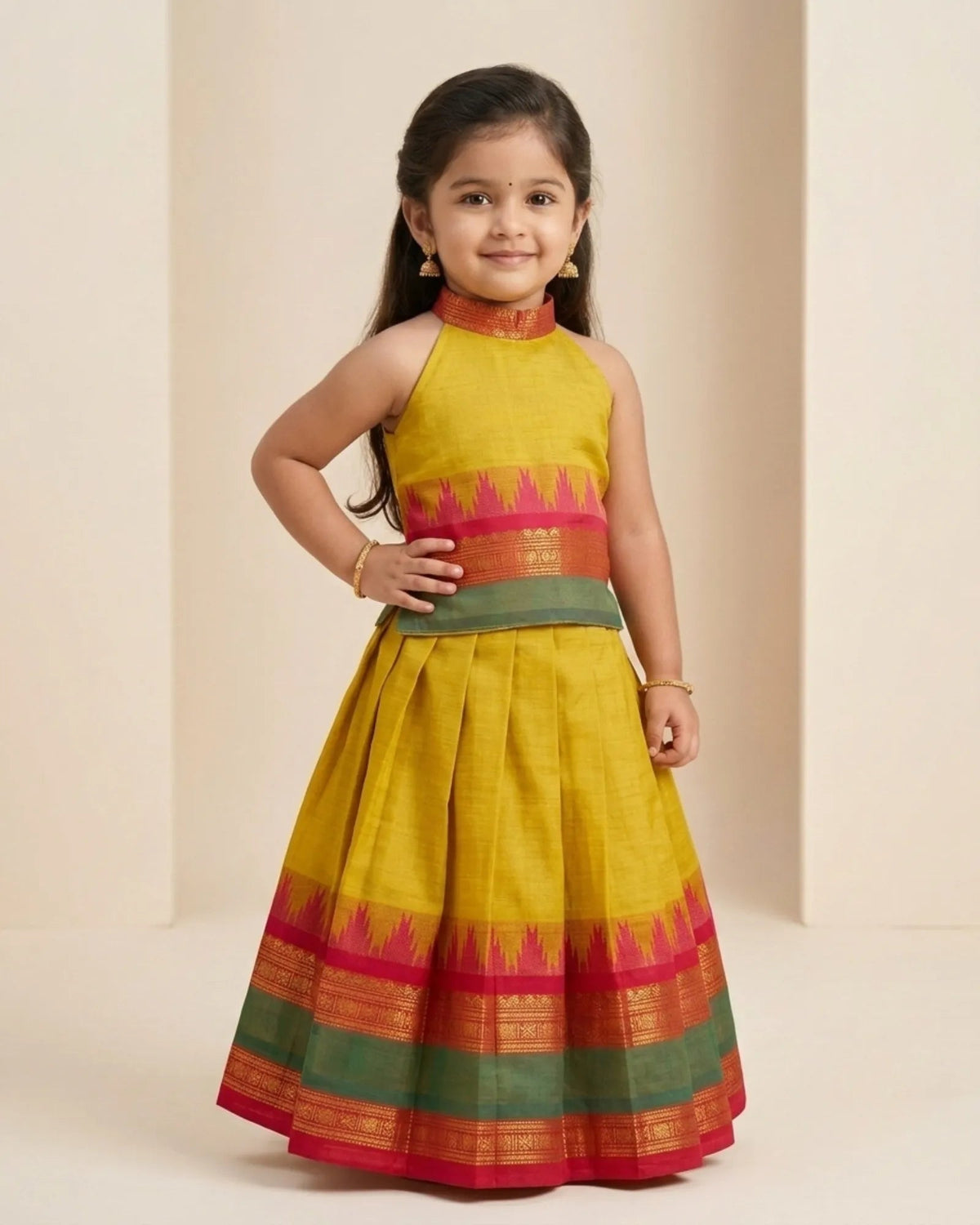 A young girl modeling a sleeveless yellow Anarkali-style dress with pink, green, and gold traditional borders, set against a warm beige studio backdrop.