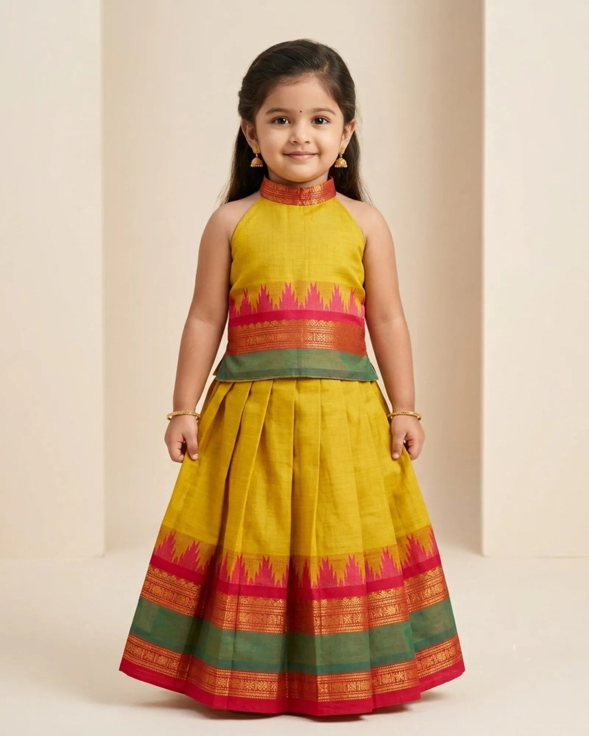 A young girl modeling a traditional Indian dress made of yellow silk with red and green patterned borders, set against a neutral beige background.