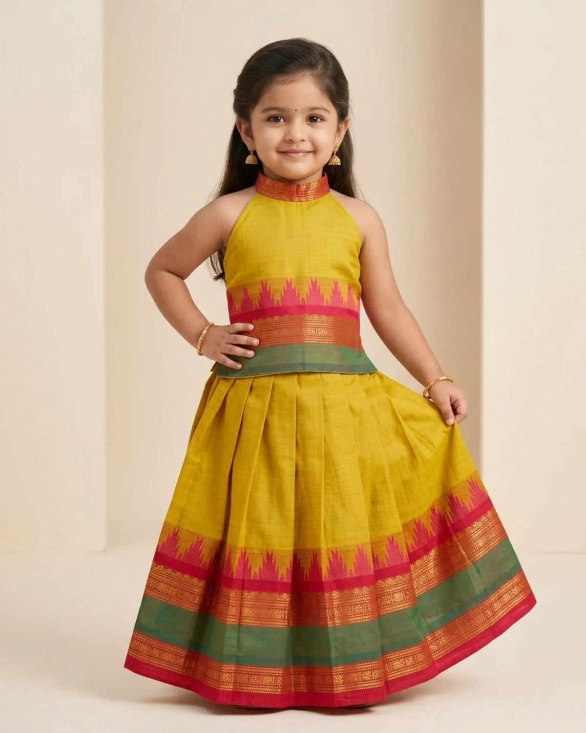 A young girl modeling a yellow cotton saree-style dress with a green and red border, standing against a neutral beige background.