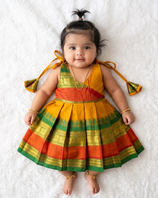 A baby girl lying on a white faux fur blanket, wearing a sleeveless pleated traditional Indian dress made of yellow, orange, and green silk with gold zari borders. She has a small black hair bun with a feather accent and is wearing gold jewelry.
