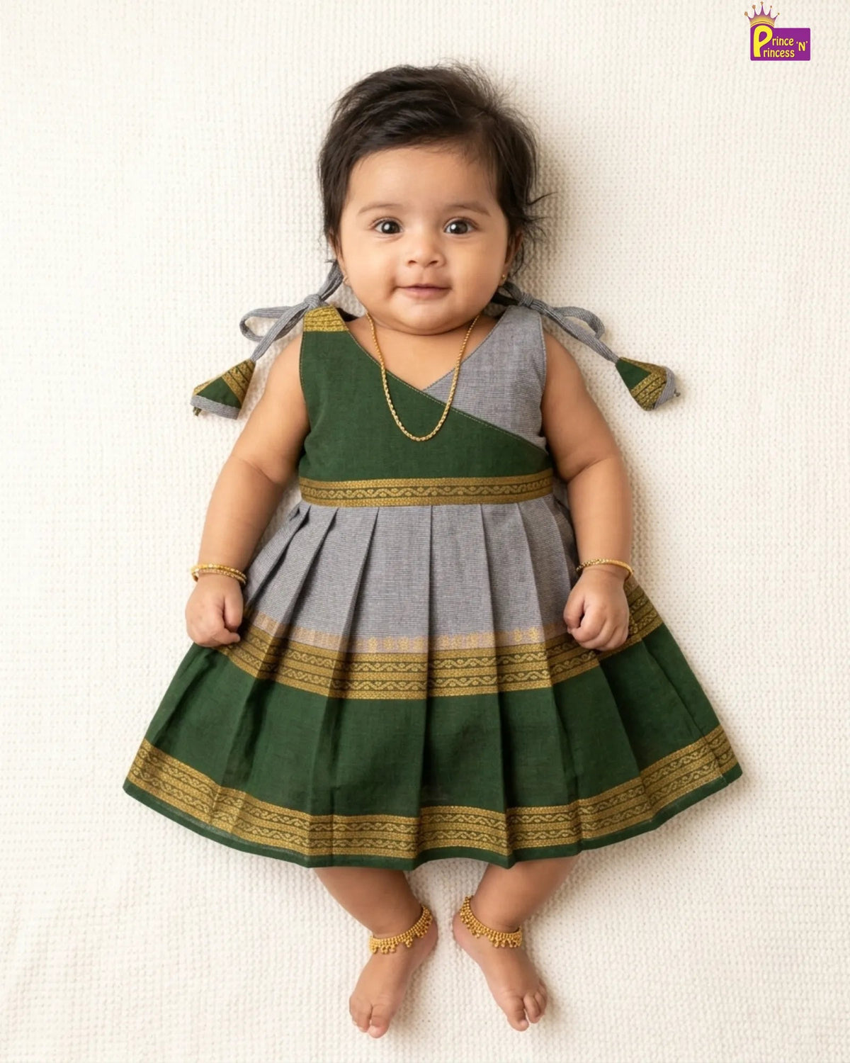 A baby girl wearing a green and gray traditional Indian dress with gold borders, lying on a textured cream background. She is wearing gold jewelry including a necklace, bangles, and ankle bracelets.