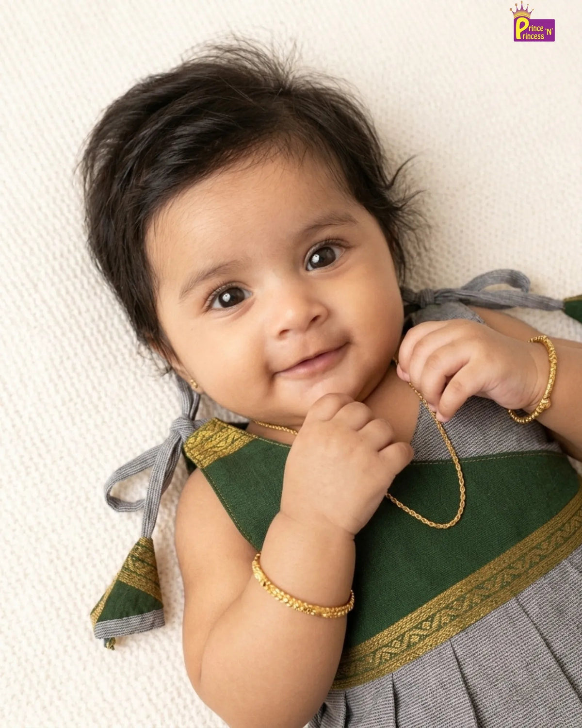 A baby wearing a green and gray traditional outfit with gold jewelry, lying on a textured white blanket.
