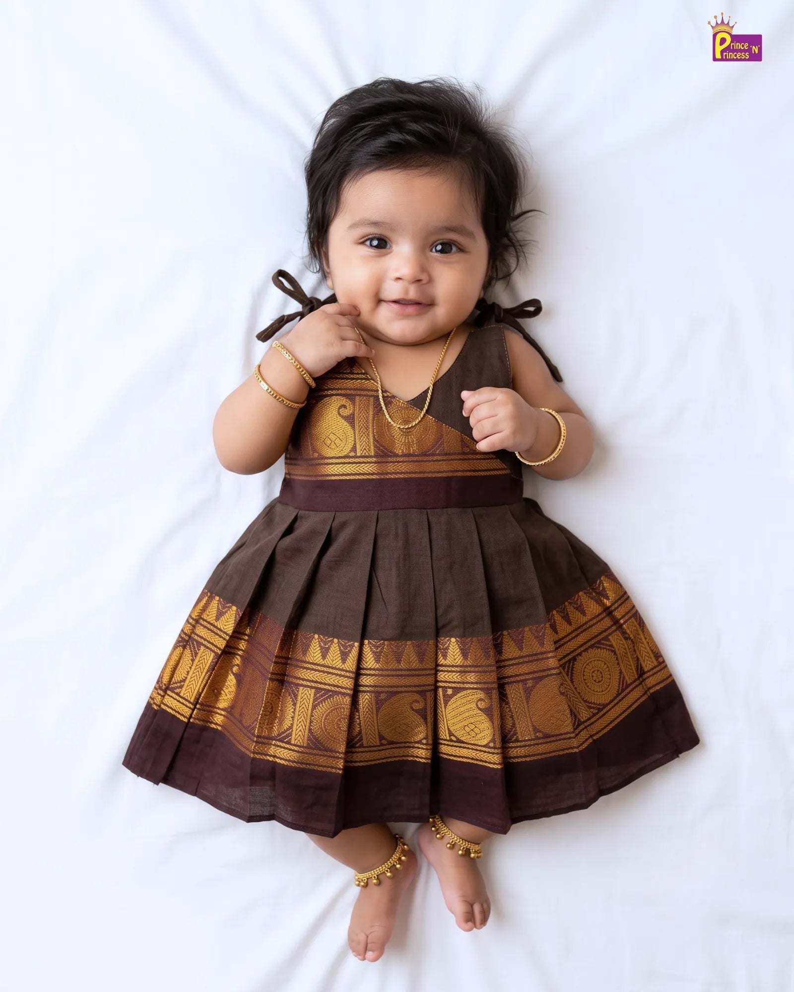 A baby girl lying on a white sheet wearing a brown and gold traditional dress with pleated skirt and gold jewelry.