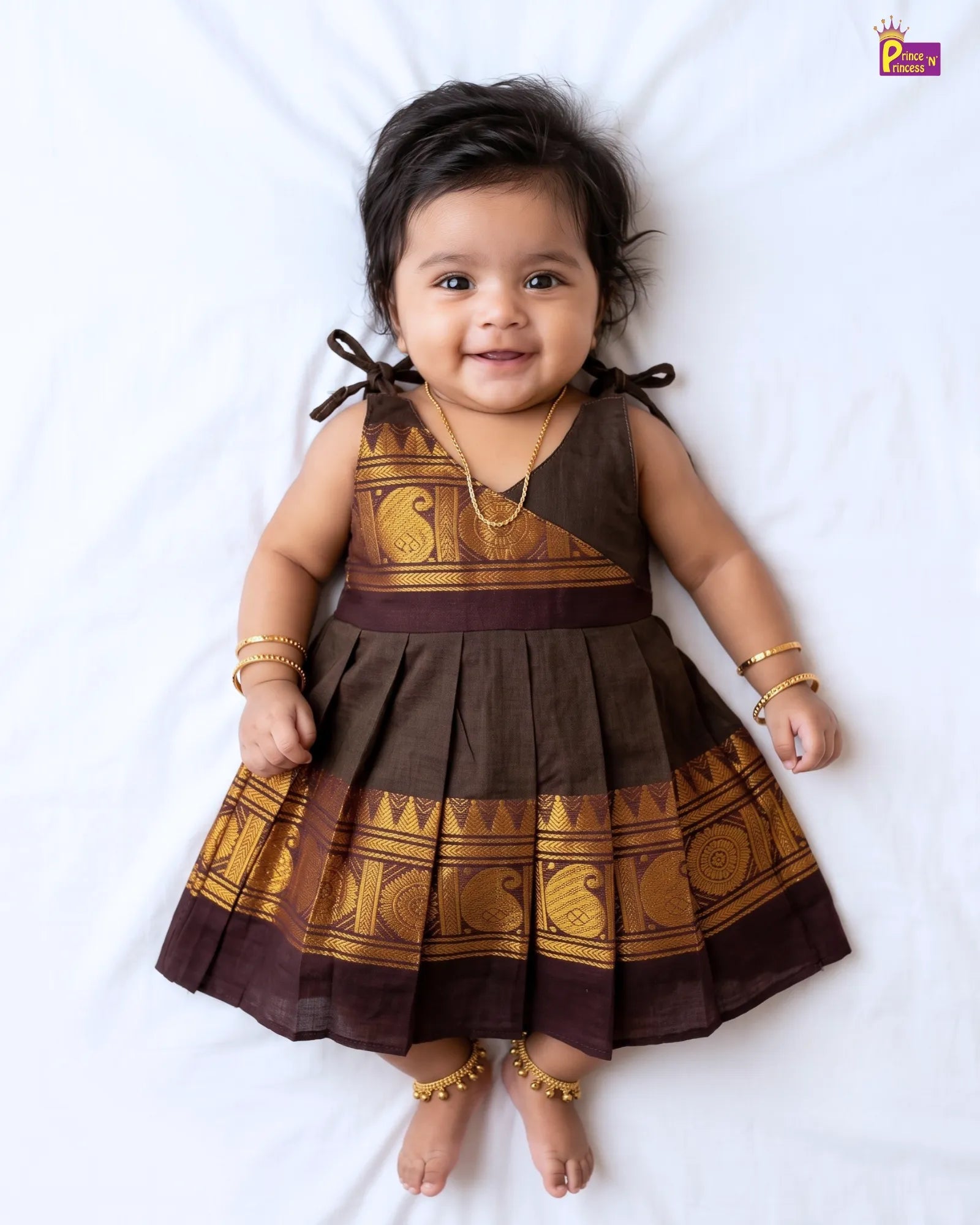 A baby girl lying on a white sheet wearing a brown and gold traditional sleeveless dress with pleated skirt, gold jewelry, and gold anklets.