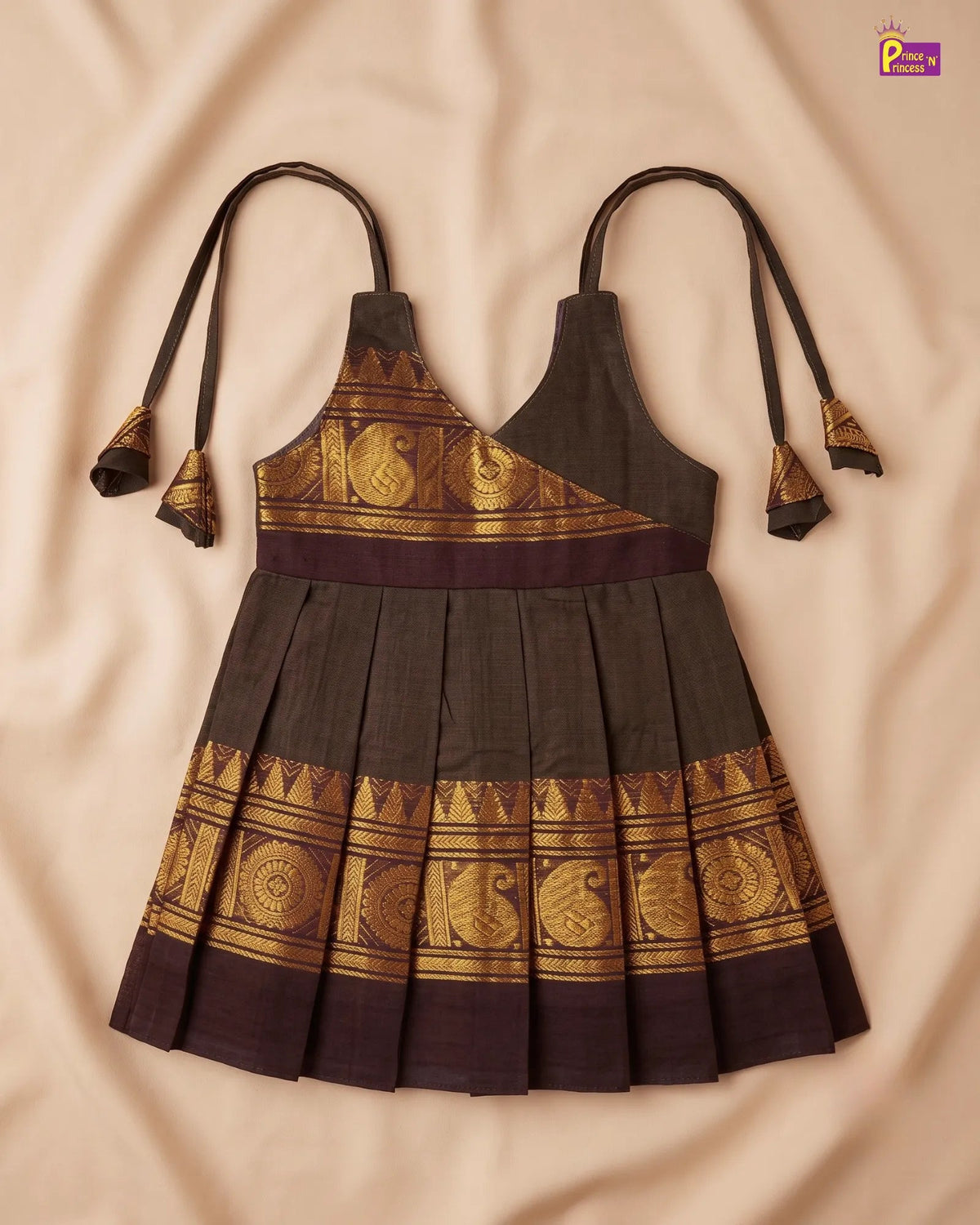 A sleeveless dark brown and gold printed halter dress laid flat on a beige fabric background. The dress features a pleated skirt, a wide gold patterned band, and matching tie straps with decorative gold tassels.