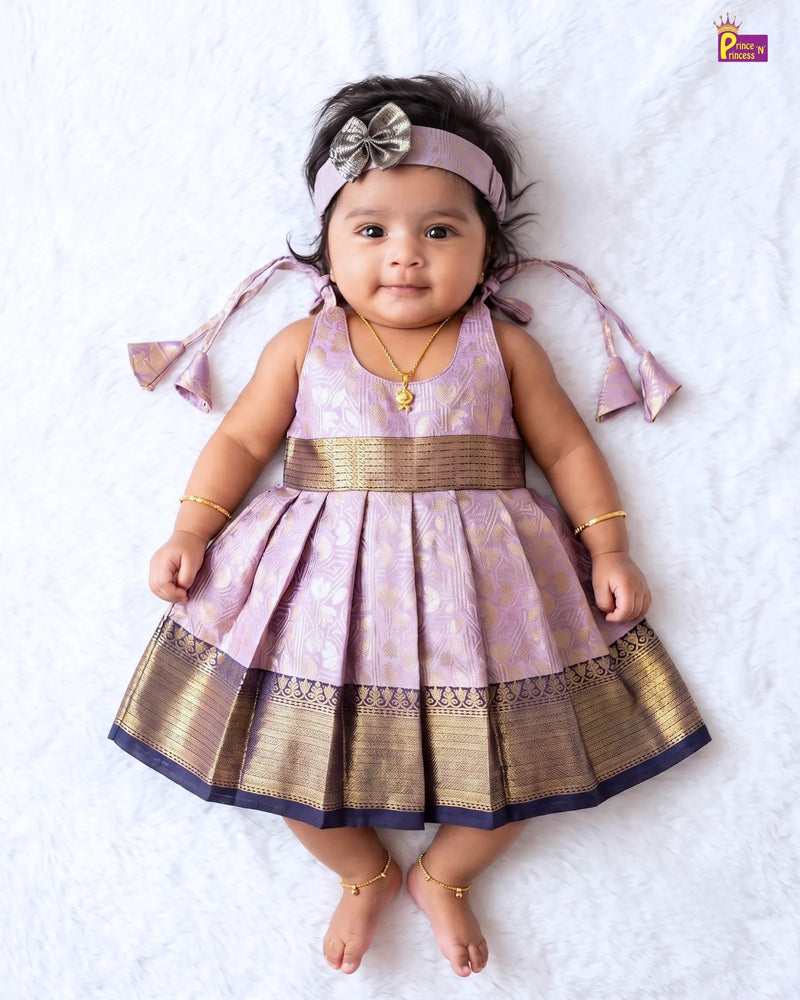 A baby girl lying on a white textured blanket, wearing a lavender and gold traditional Indian dress with a matching headband and gold jewelry.