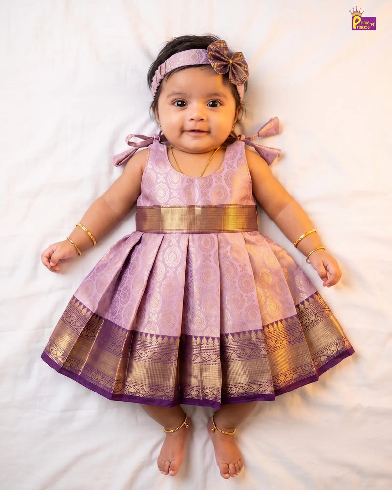 A baby girl lying on a white sheet wearing a lavender and gold traditional Indian dress with a pleated skirt, a wide gold waist sash, a matching lavender bow headband, and gold bangles and ankle bracelets.