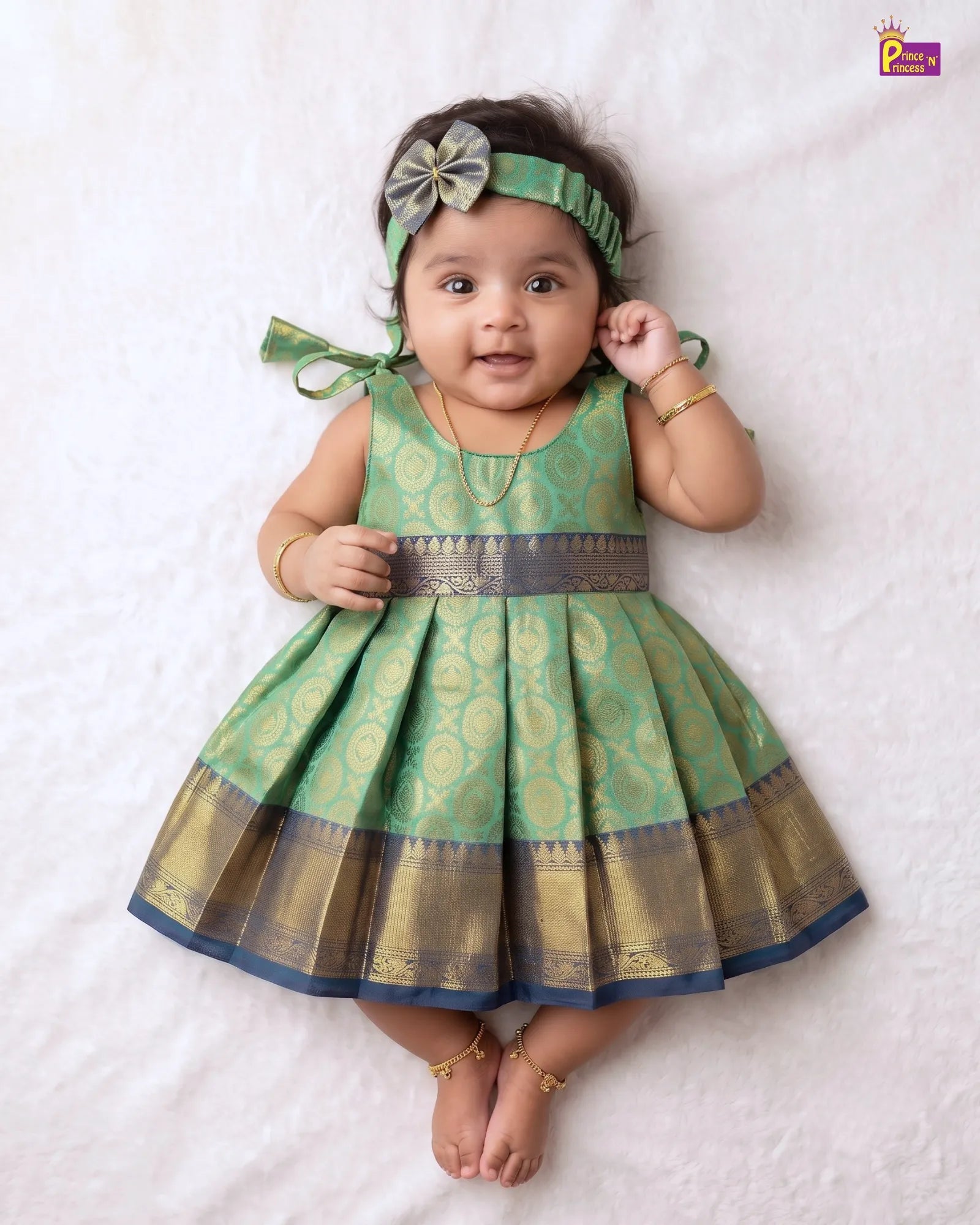 A baby girl wearing a green and gold patterned dress with a matching headband, lying on a white textured blanket.