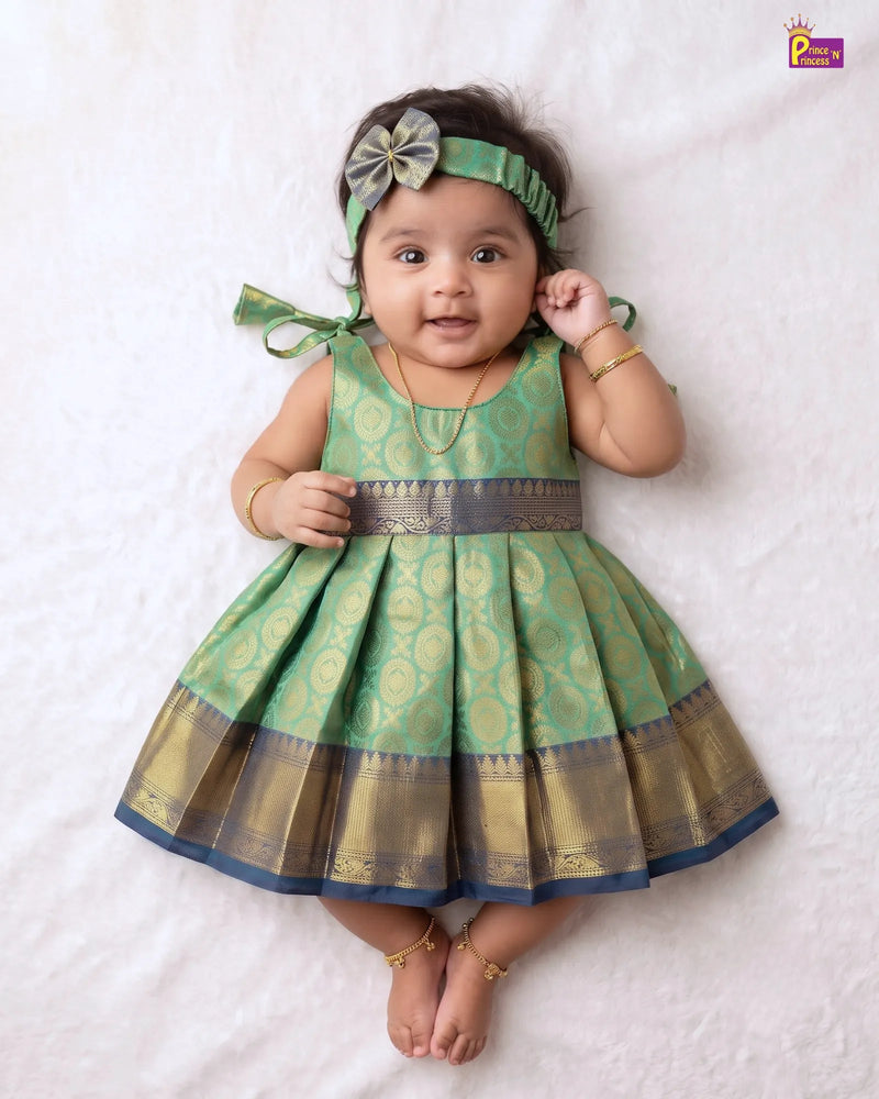 A baby girl wearing a green and gold patterned dress with a matching headband, lying on a white textured blanket.