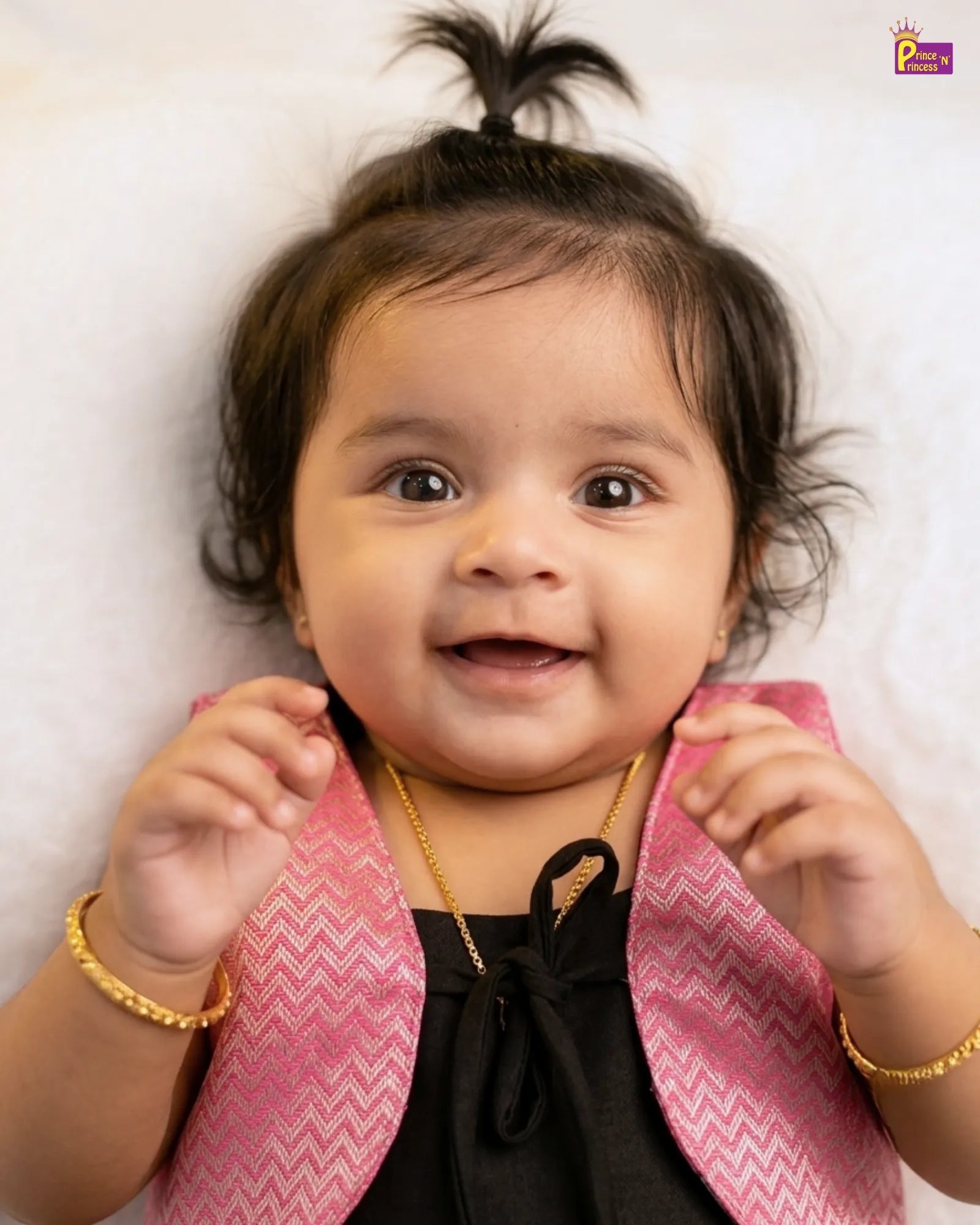 Baby wearing a pink jacket over a black outfit with gold jewelry on a white background