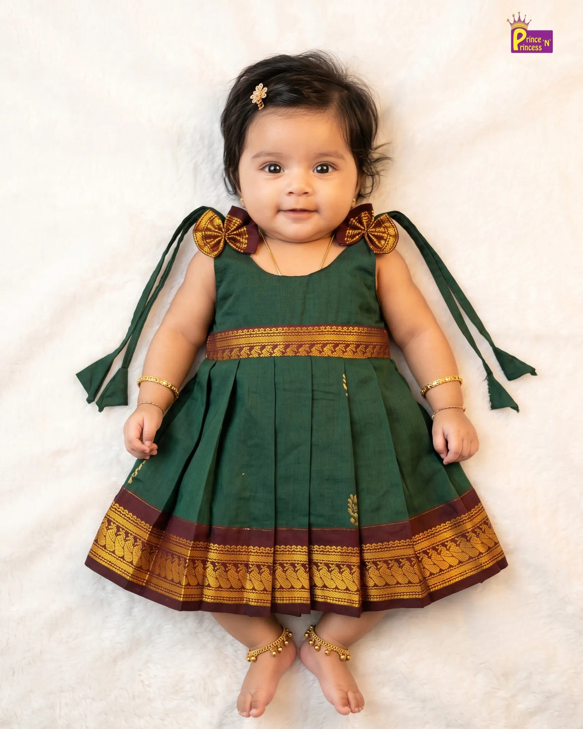 A baby girl wearing a dark green traditional dress with gold and maroon borders, lying on a white textured blanket. The dress has bow details on the shoulders with long green ribbons and gold trim.
