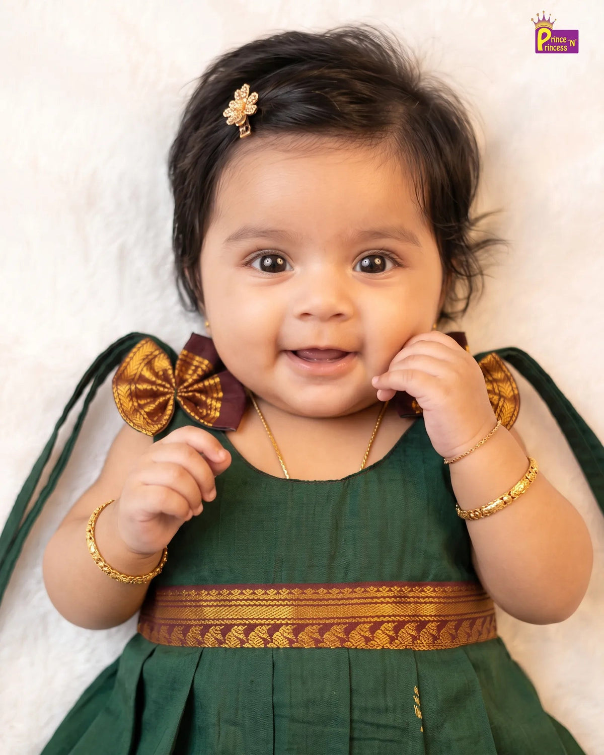 A smiling baby girl wearing a dark green traditional dress with gold and maroon embroidery, lying on a white textured blanket. She has a gold hair clip and gold bracelets.
