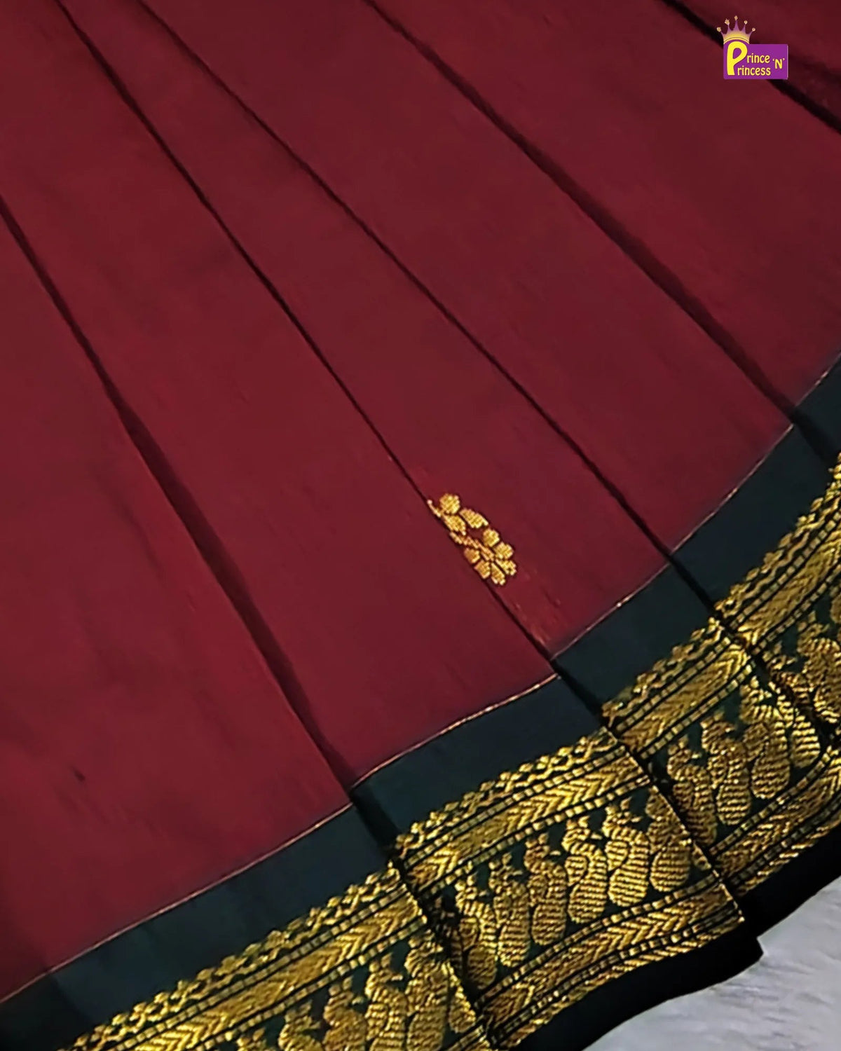 A close-up of a deep red fabric with pleated texture, featuring a decorative gold floral motif and a wide black and gold embroidered border.