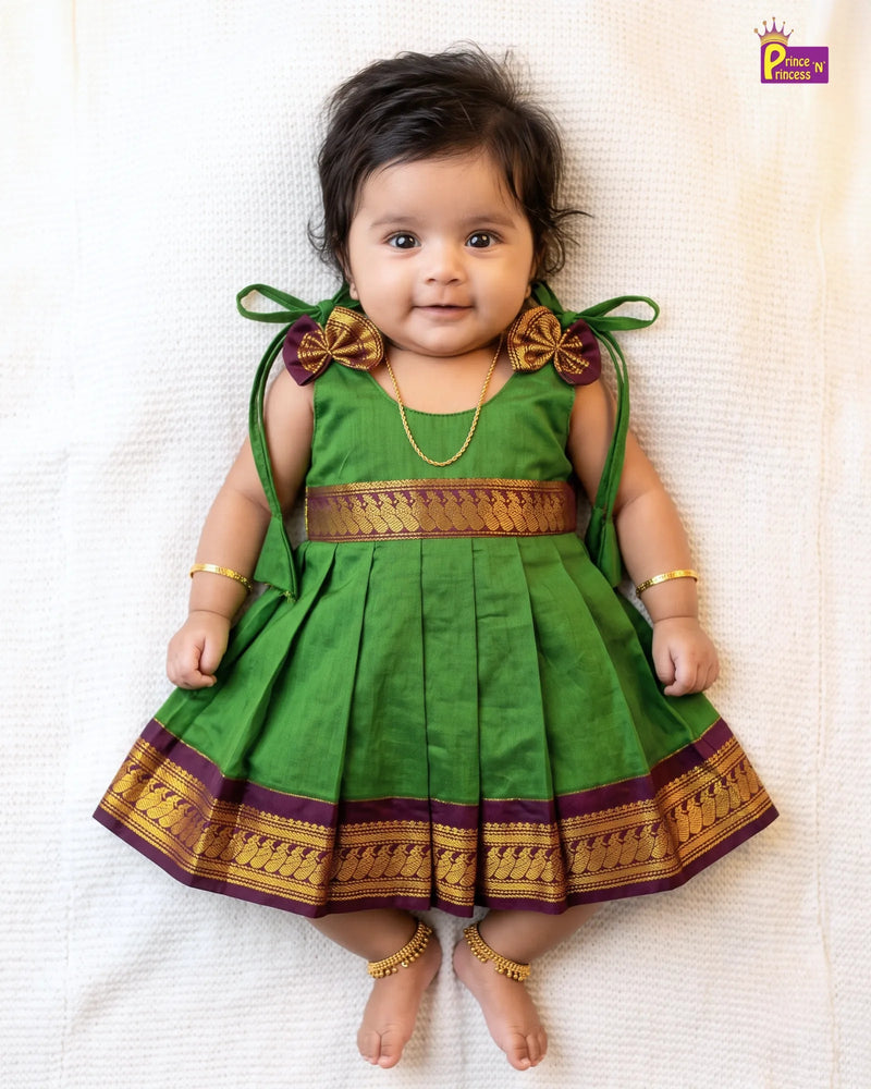 A baby girl lying on a textured white blanket, wearing a green traditional Indian dress with gold and purple borders, bow details on the shoulders, and gold jewelry.