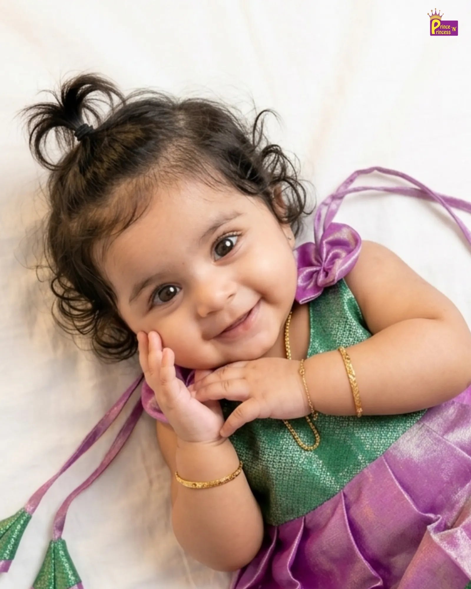 Child wearing a green and purple traditional outfit with a gold bracelet, lying on a white background.