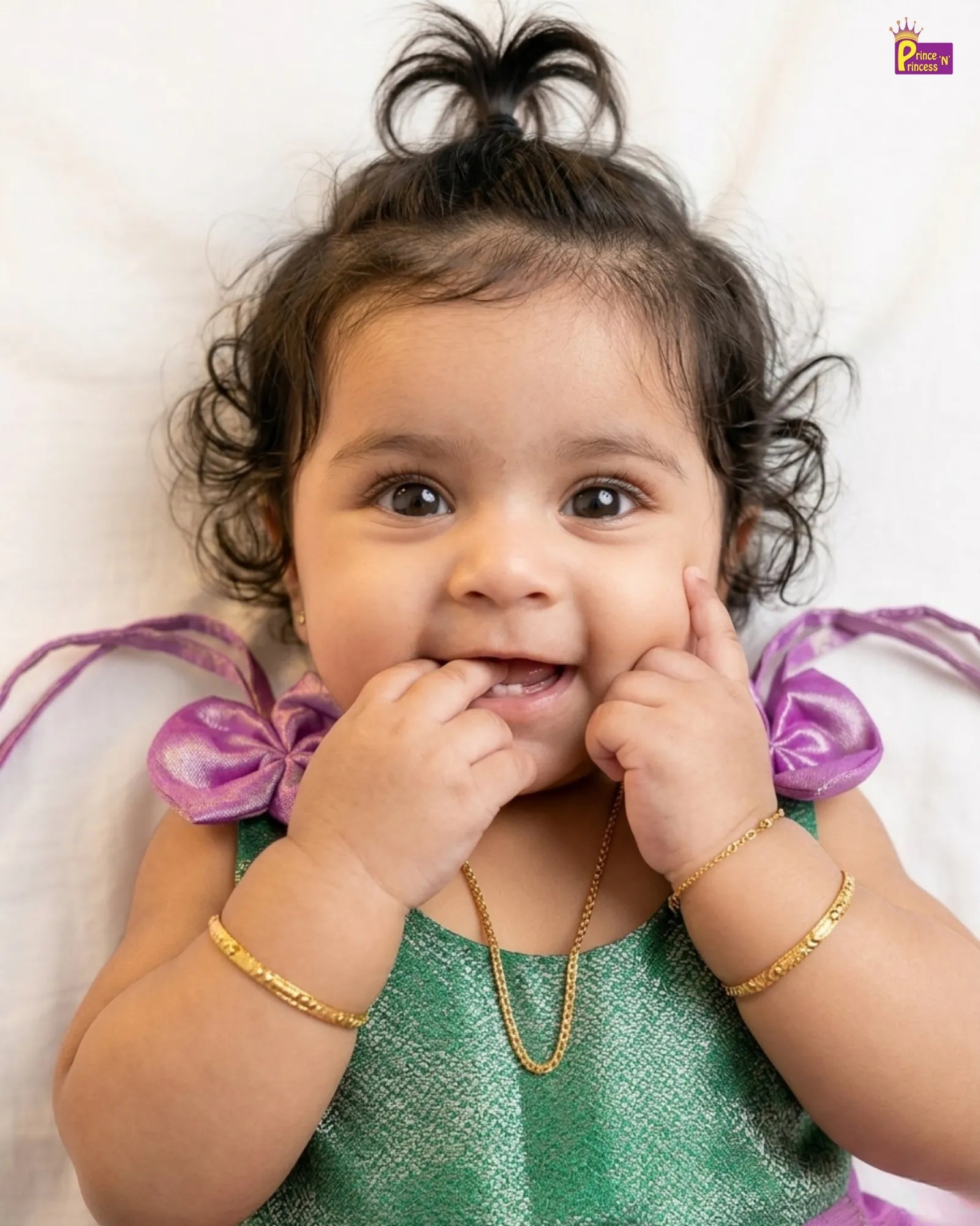Baby wearing a green outfit with purple accents and gold jewelry on a white background
