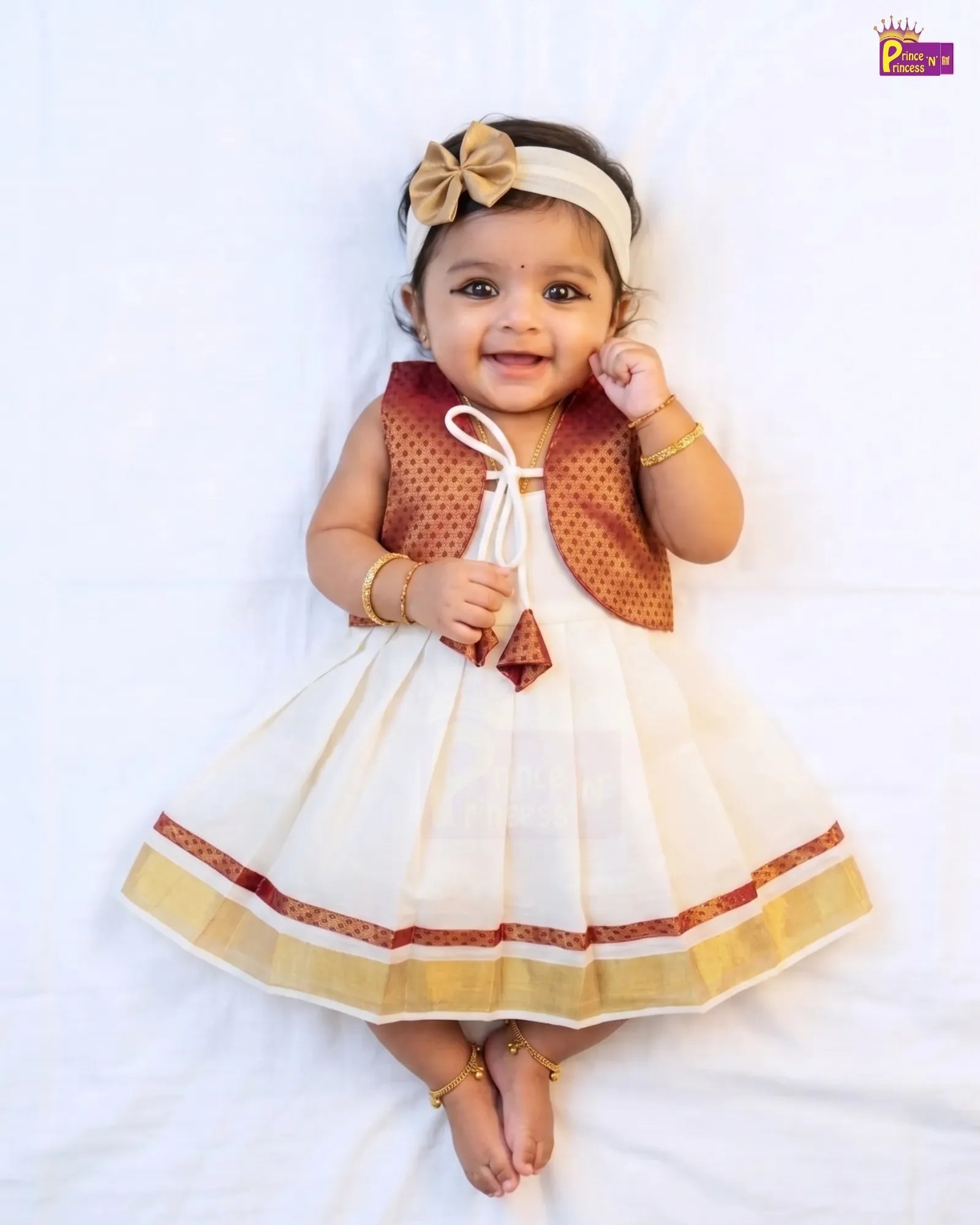A baby girl lying on a white sheet wearing a traditional Indian dress with a white body, a rust-orange patterned sleeveless overlay, and a matching headband with a beige bow.