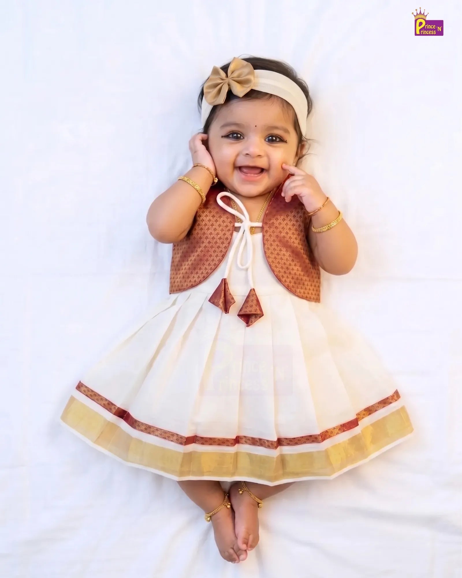 A baby girl lying on a white sheet wearing a cream and brown traditional Indian dress with a matching headband.