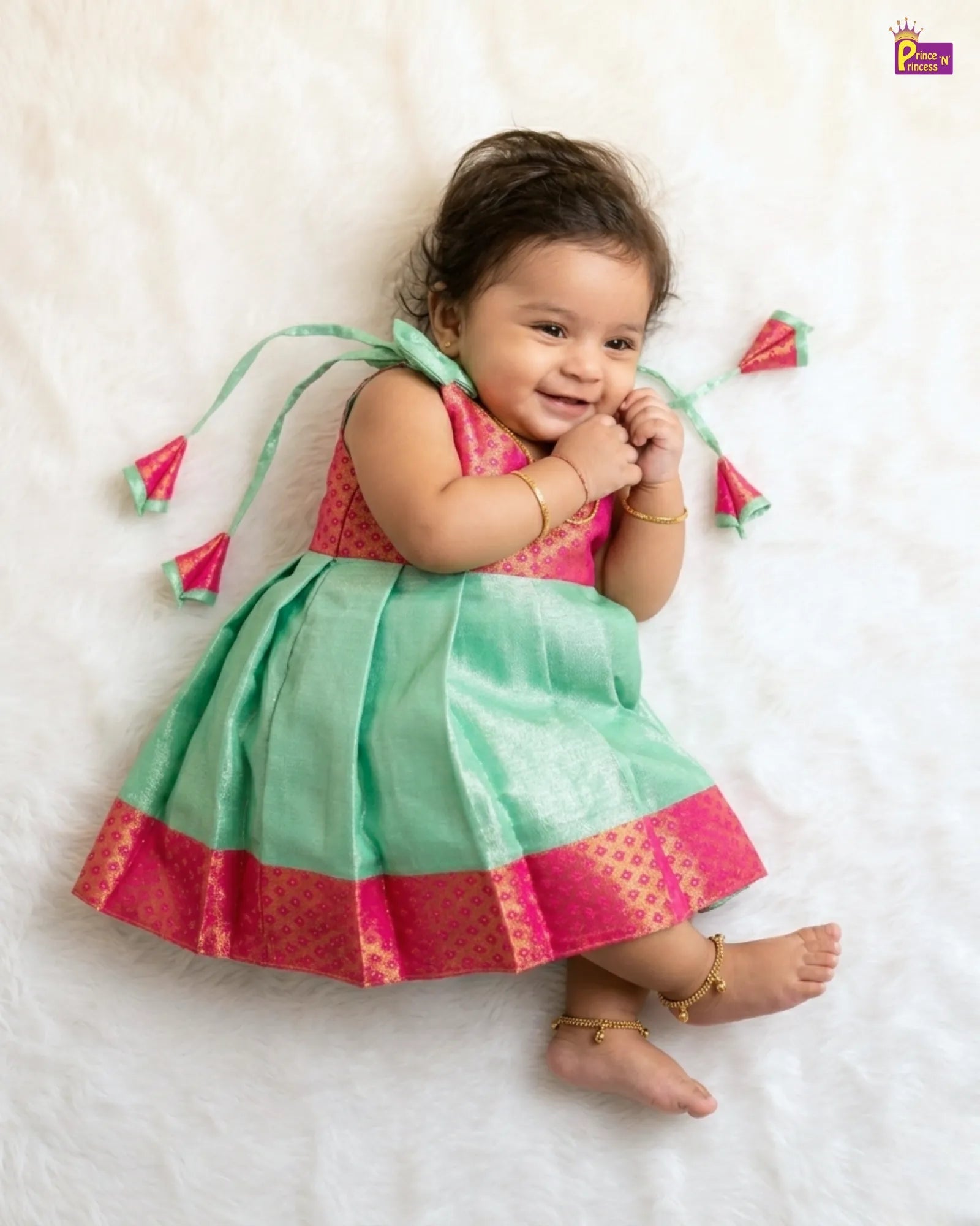 Baby wearing a pink and green dress with tassels on a white background