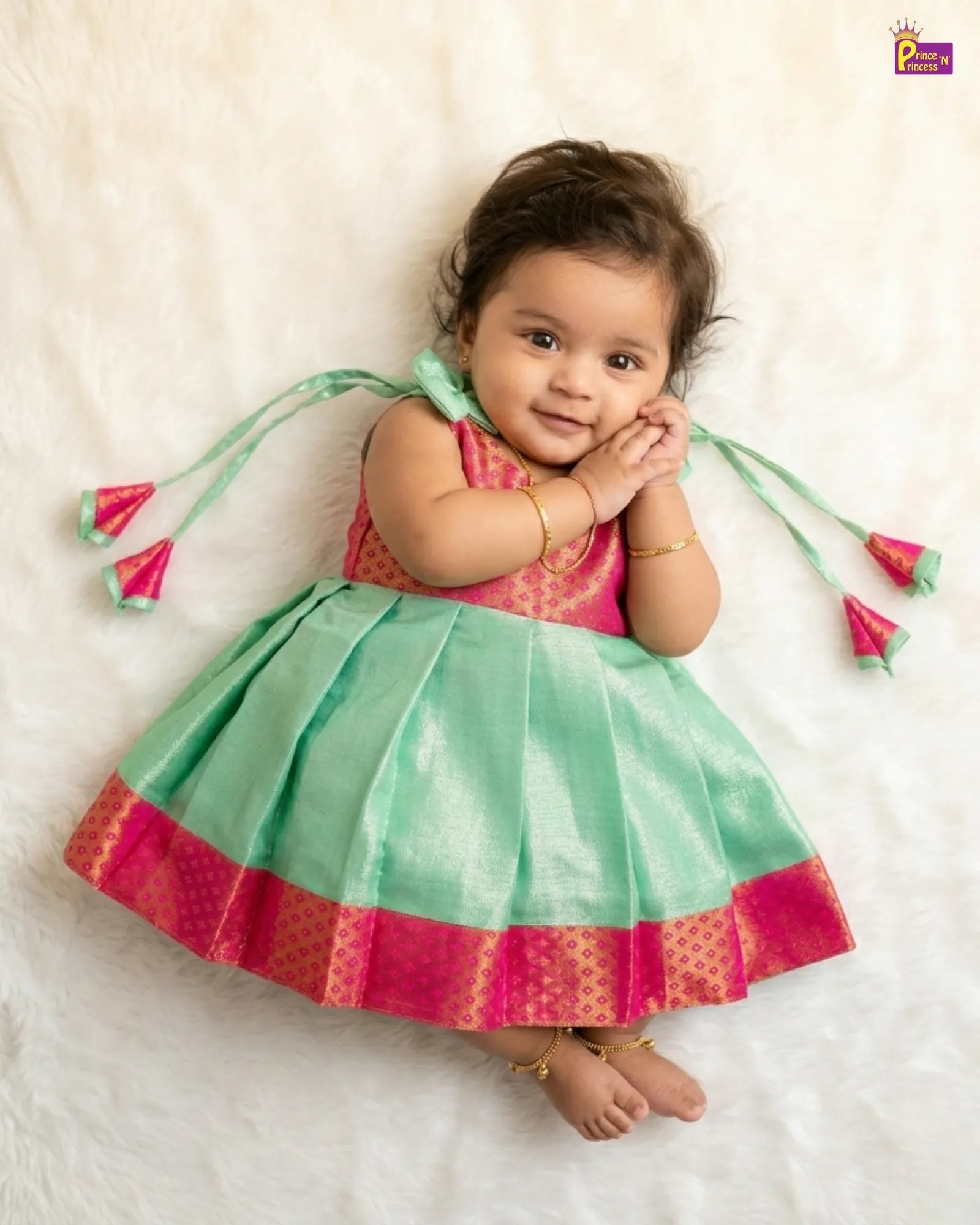 Baby wearing a green and pink traditional outfit with tassels on a white background