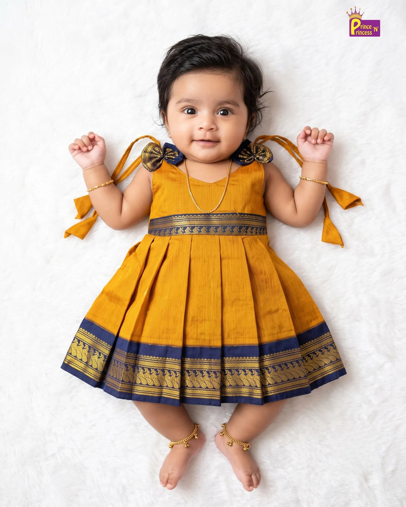 A baby girl lying on a white textured blanket, wearing a mustard yellow traditional dress with navy blue and gold borders, bow details on the shoulders, and matching ribbon ties. She is wearing gold jewelry including a necklace, bracelets, and ankle chains.